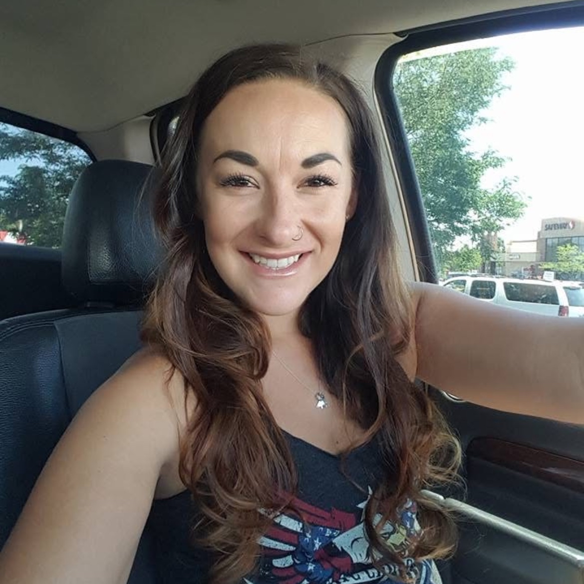Smiling woman with long brown hair, wearing a black tank top with a patriotic design, sitting inside a car, with trees and parked vehicles visible outside.