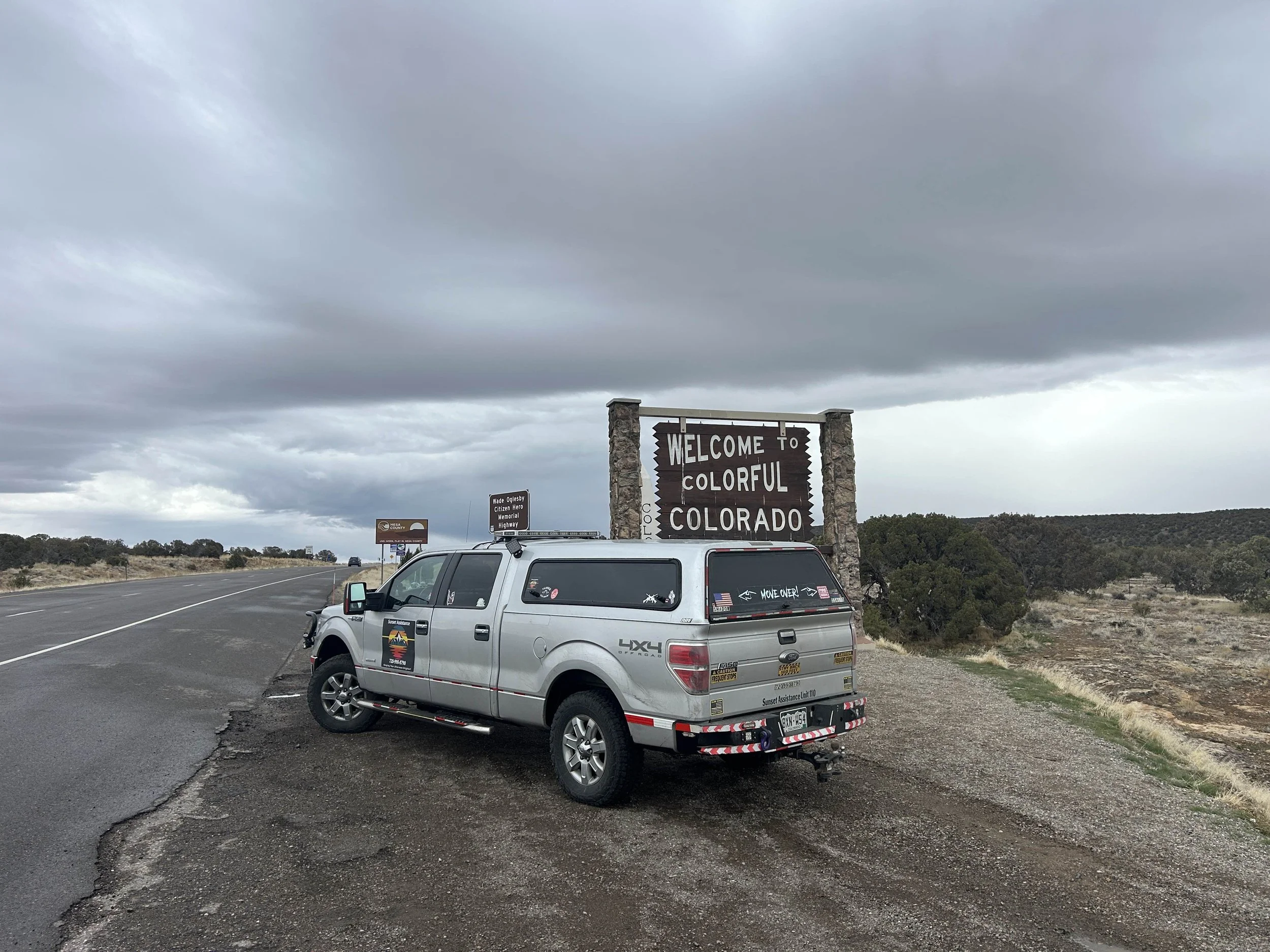A silver pickup truck with sunset assistance decals parked at the roadside next to a welcome sign that says 'Welcome to Colorado'. The sky is overcast with gray clouds, and there are several highway signs in the background.