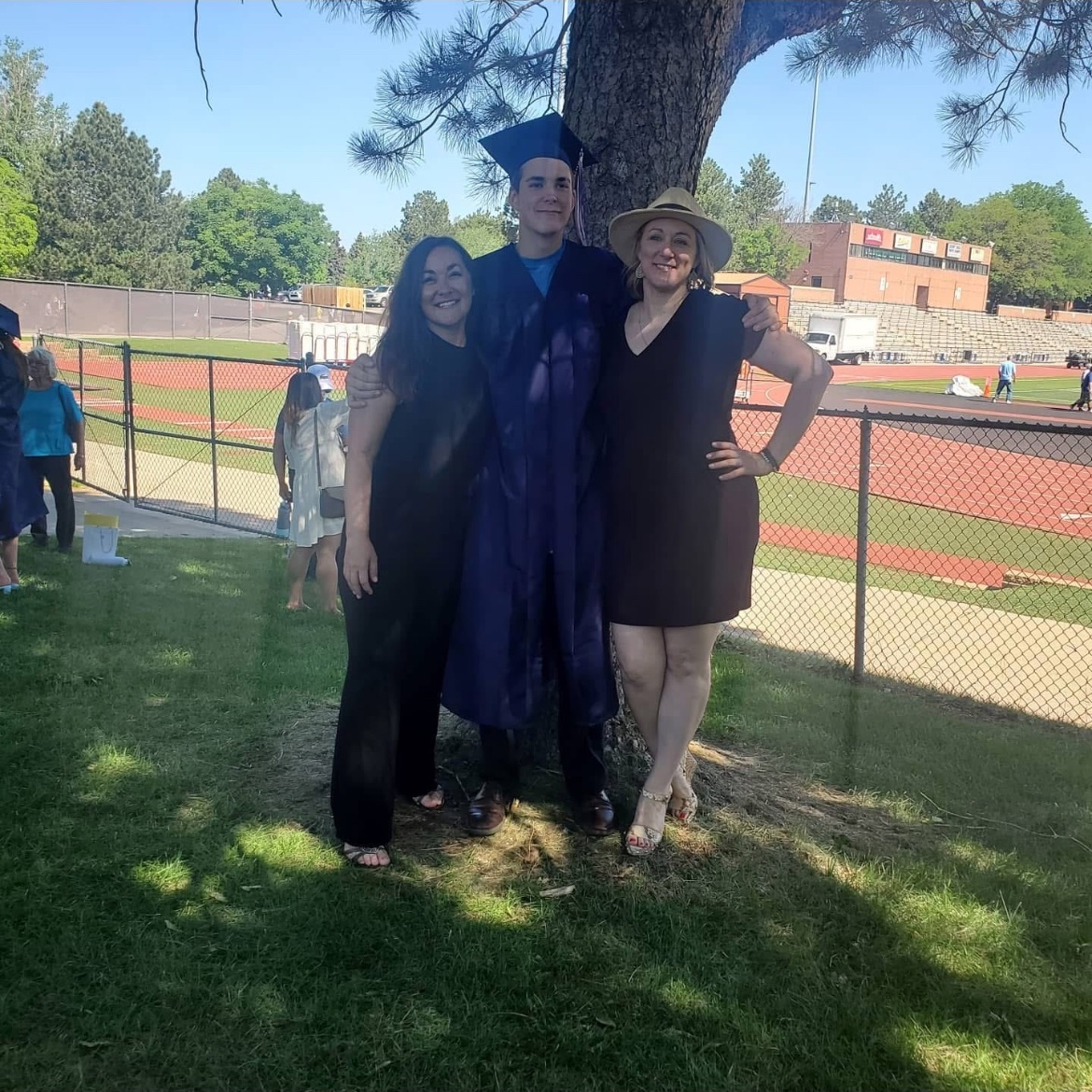 A young man in a blue graduation gown and cap is standing between two women, all smiling, outdoors near a tree with a sports field in the background. One woman is in a black dress, and the other is in a black dress with a hat.