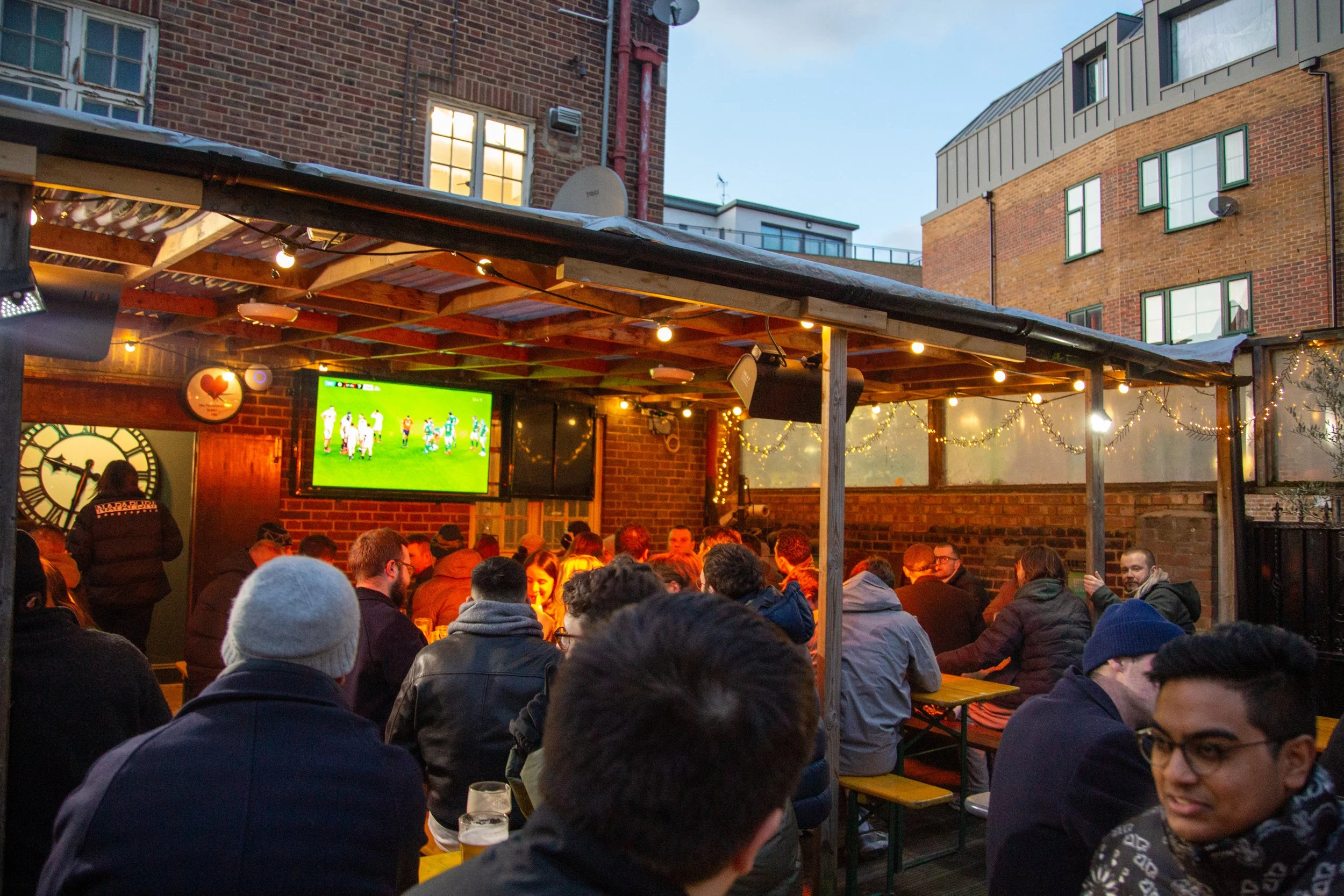 People gathered at an outdoor bar or restaurant, watching a soccer game on a large screen, with string lights and brick walls in the background in an urban setting during the evening.