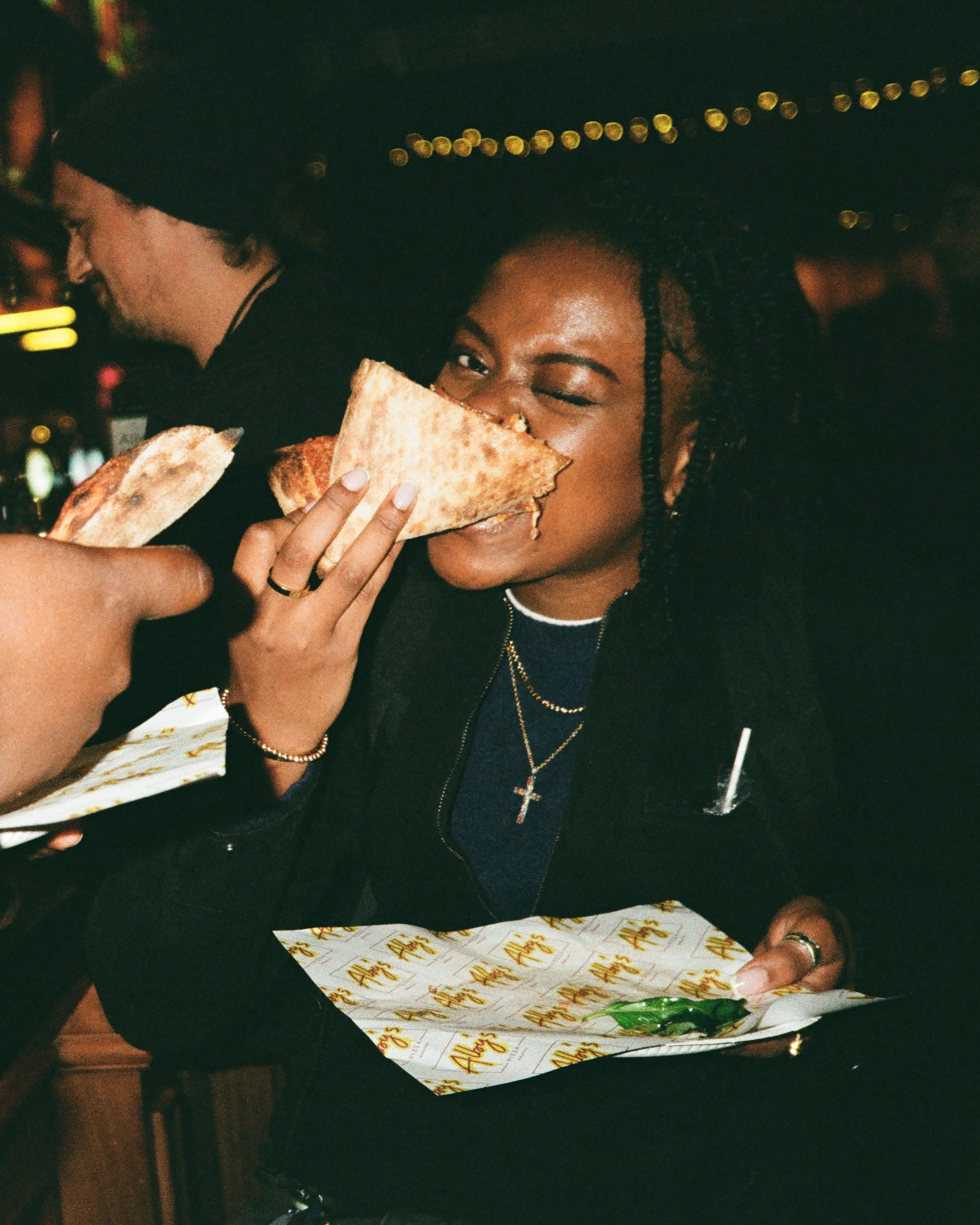Woman with braids and cross necklace eating a slice of pizza while holding a plate, in a dimly lit setting.