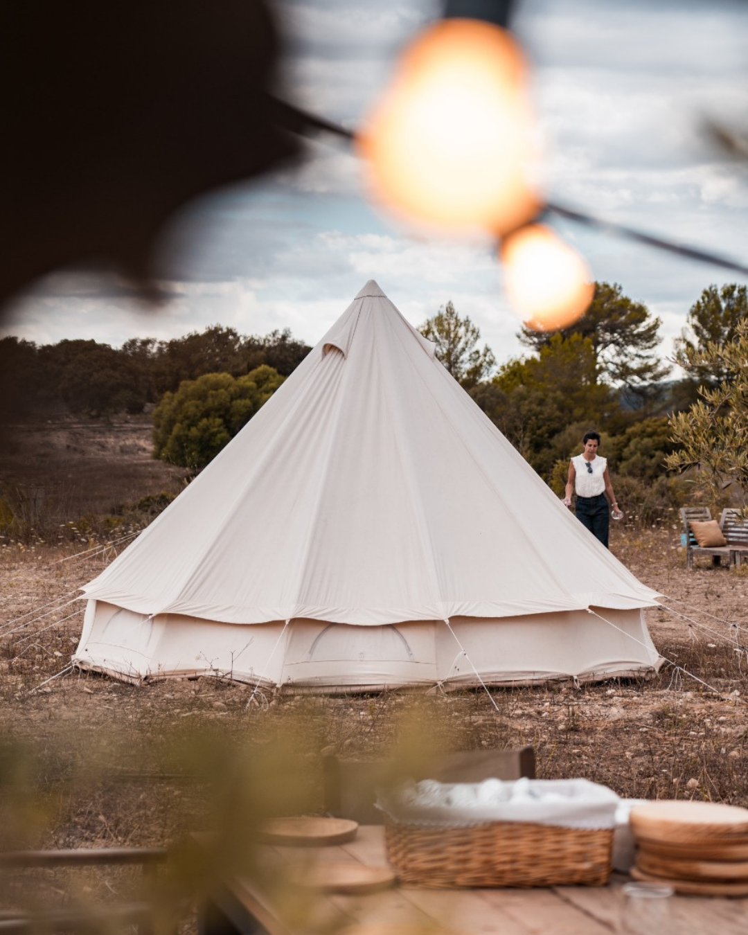 Una tienda de campaña blanca en un paisaje al aire libre con árboles y un cielo nublado, vista desde una perspectiva cercana, con unas luces decorativas en primer plano y una mujer caminando cerca de la tienda.