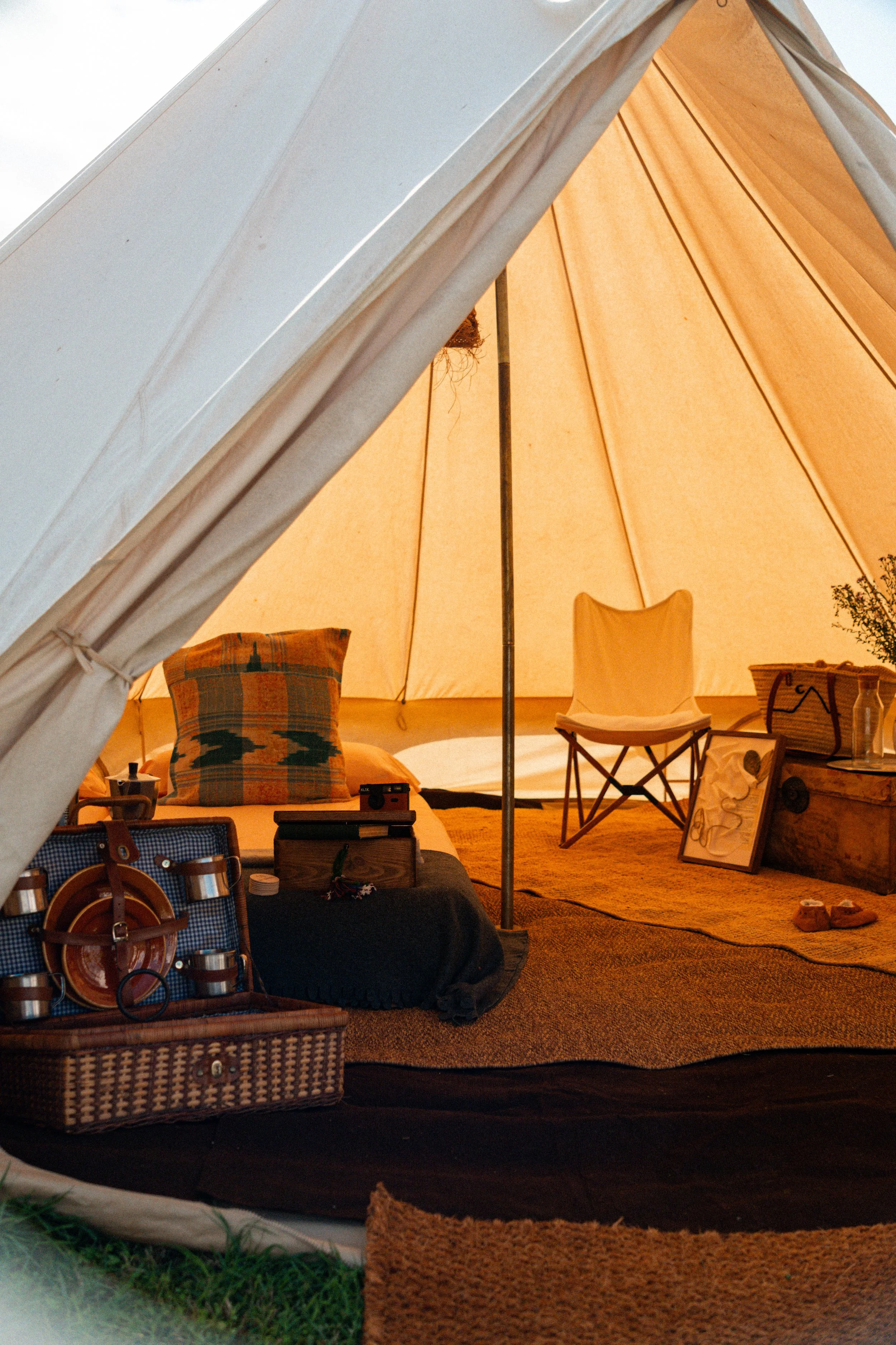 Vista desde afuera de una tienda de campaña de color beige con muebles y objetos de camping en su interior, incluyendo una silla, una bolsa, y utensilios de cocina, en un ambiente de camping.