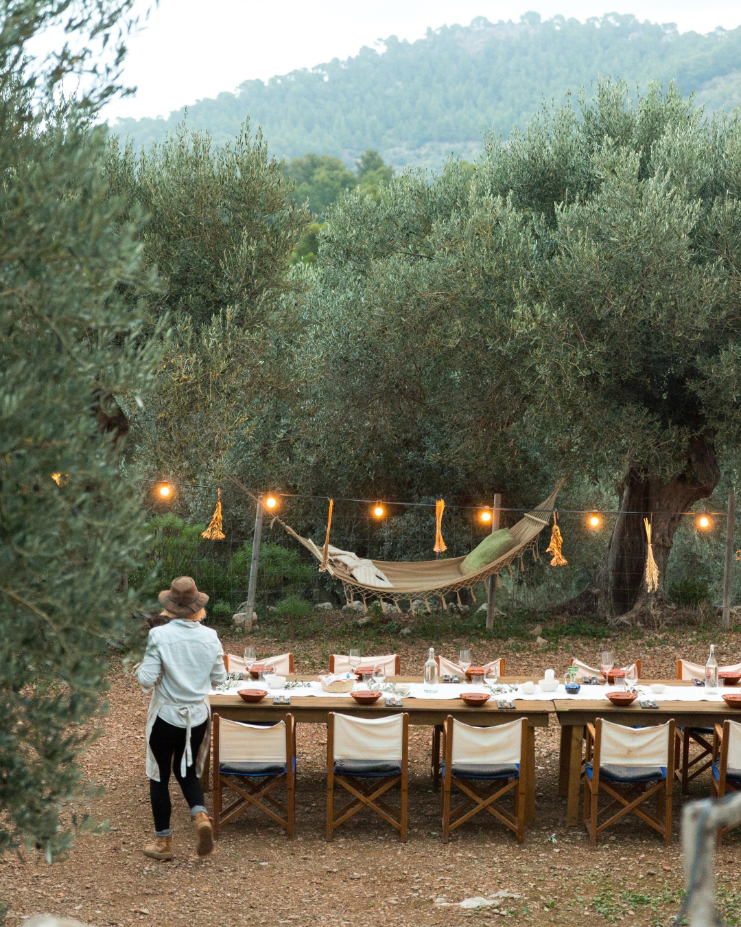 Una mesa larga para comer en un espacio al aire libre, decorada con platos, copas y botellas, rodeada de sillas blancas. En el fondo, hay un árbol grande con luces y una hamaca colgada entre las ramas, con la naturaleza y colinas en el horizonte.