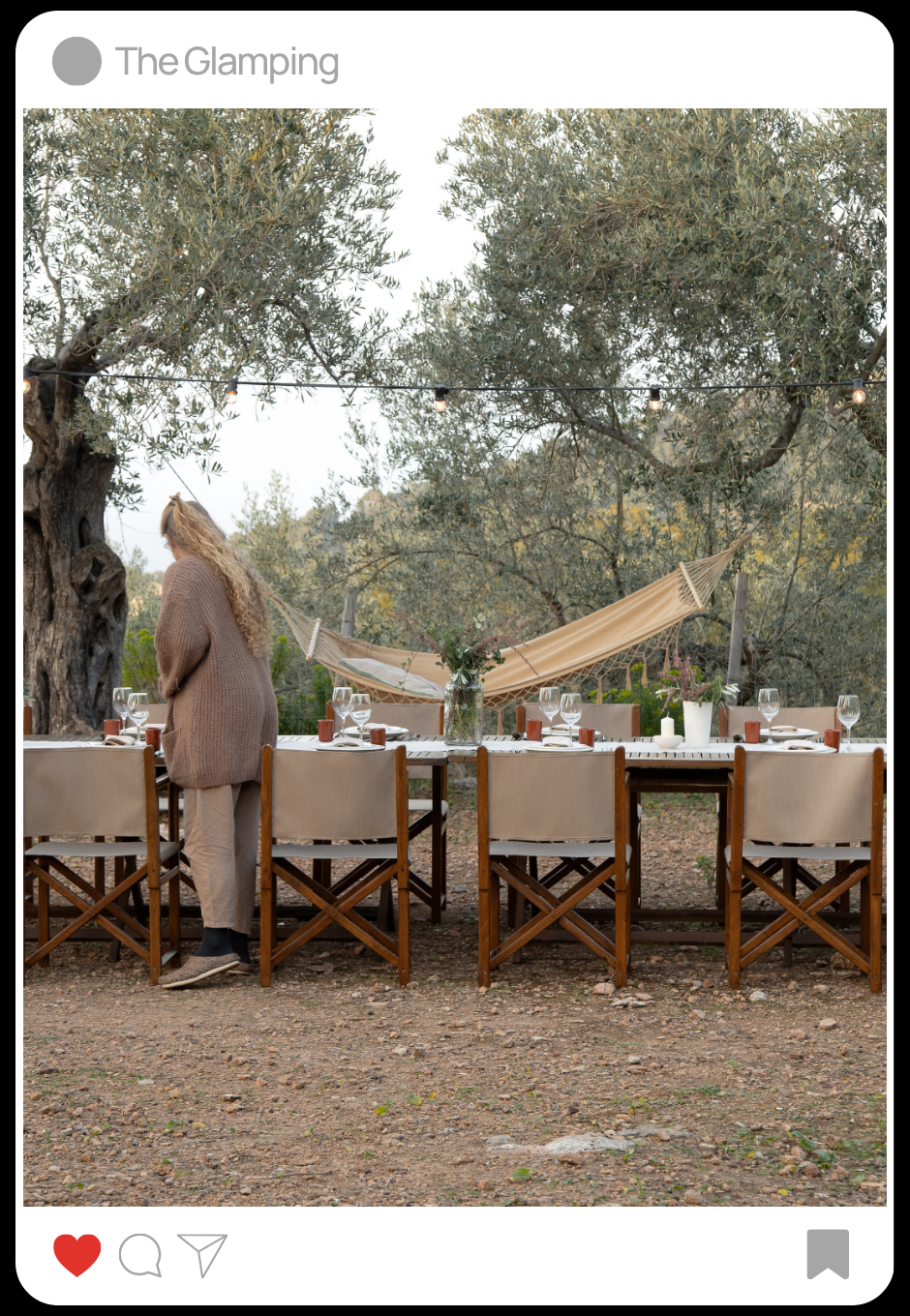 Una mesa de comedor al aire libre con sillas de madera y un mantel blanco, decorada con jarrones de flores y velas, en un entorno natural con árboles y una hamaca colgada entre ellos.