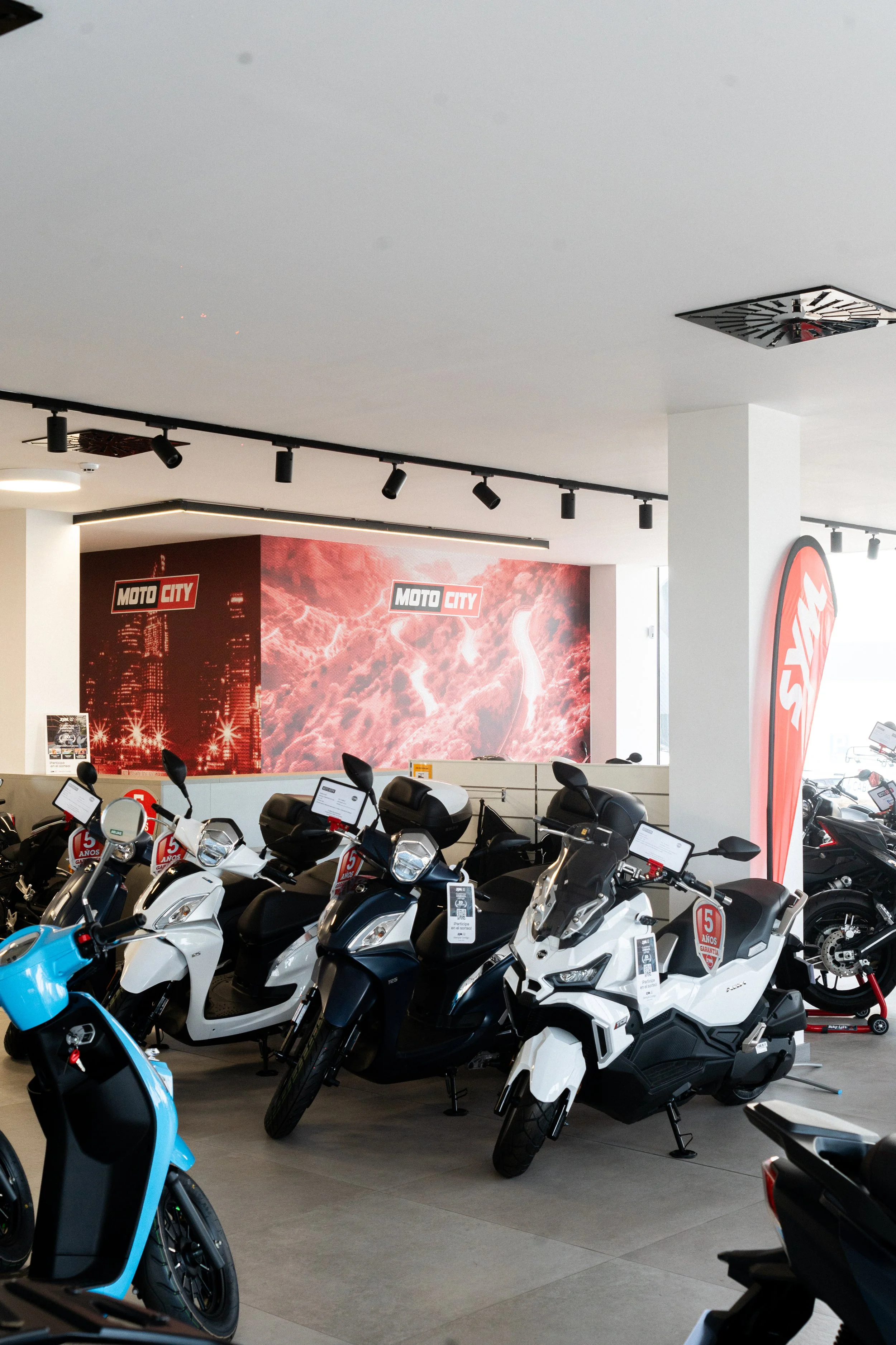 Varios scooters de motoshop en exhibición en un showroom, con una publicidad de Moto City y una bandera con el logo en el fondo.