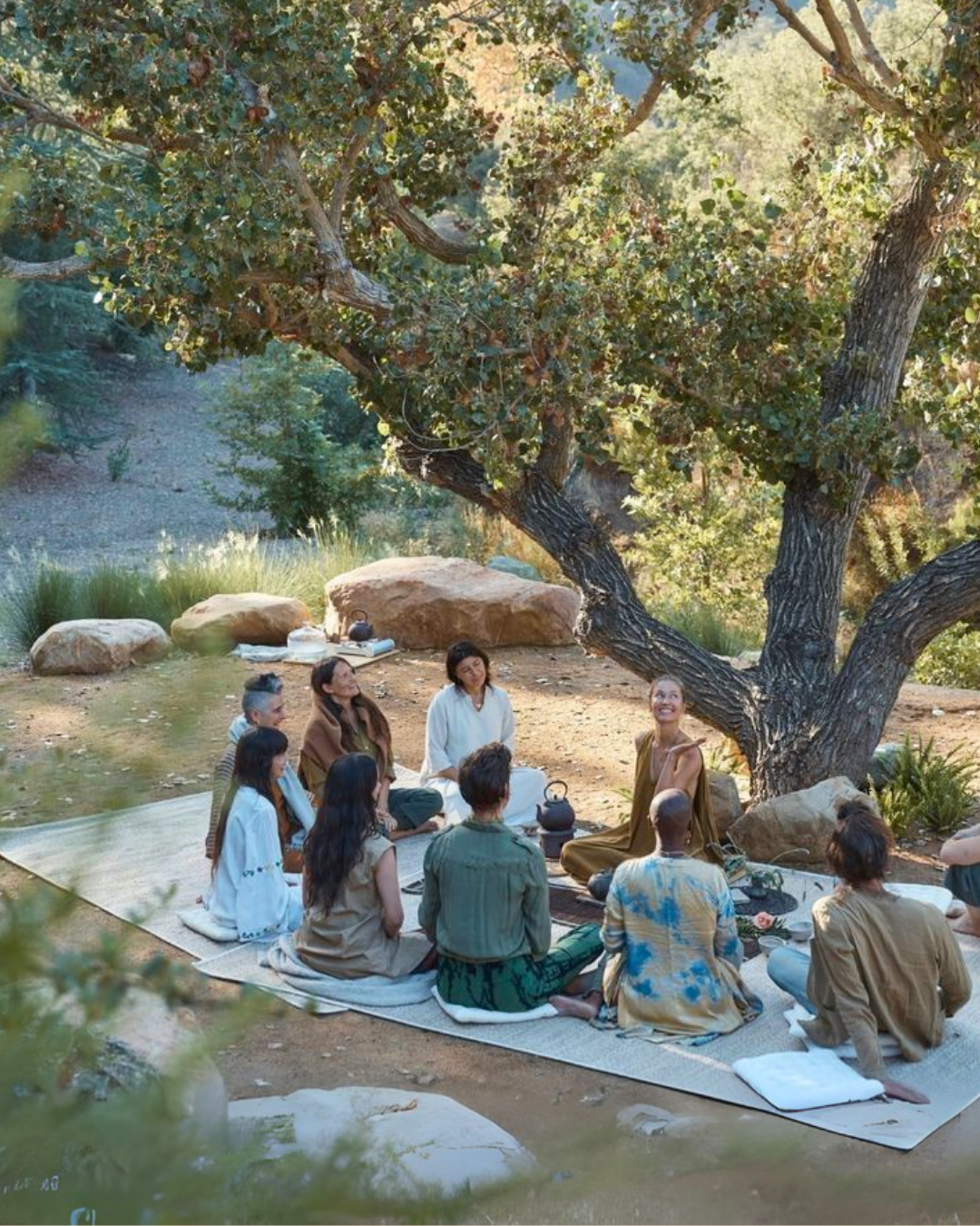 Grupo de personas sentadas en un tapete al aire libre debajo de un árbol grande, participando en una ceremonia o reunión. Hay una mujer en el centro que parece estar liderando la actividad, rodeada de otras personas que escuchan atentamente.