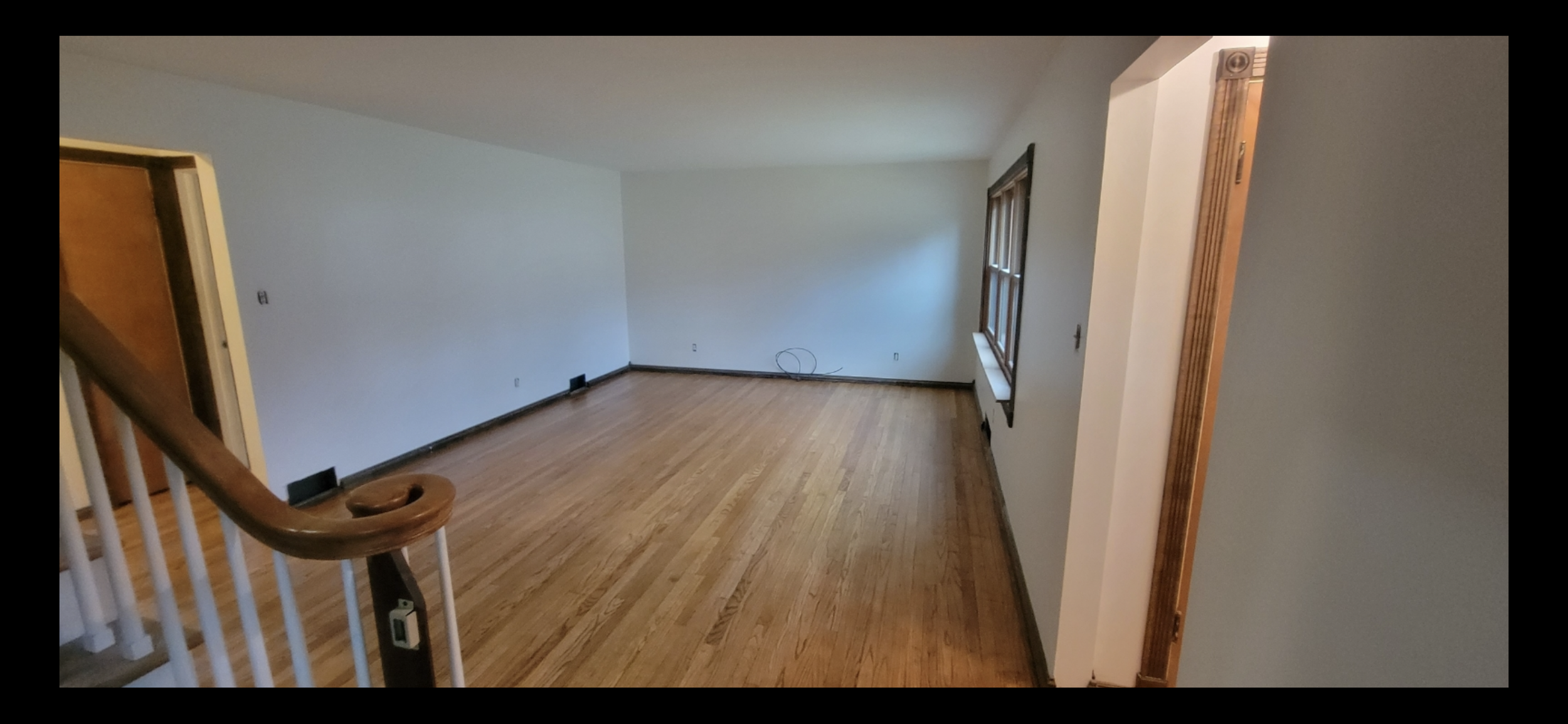 Empty living room with hardwood floors, white walls, a window with wooden trim, and a staircase railing in the foreground.
