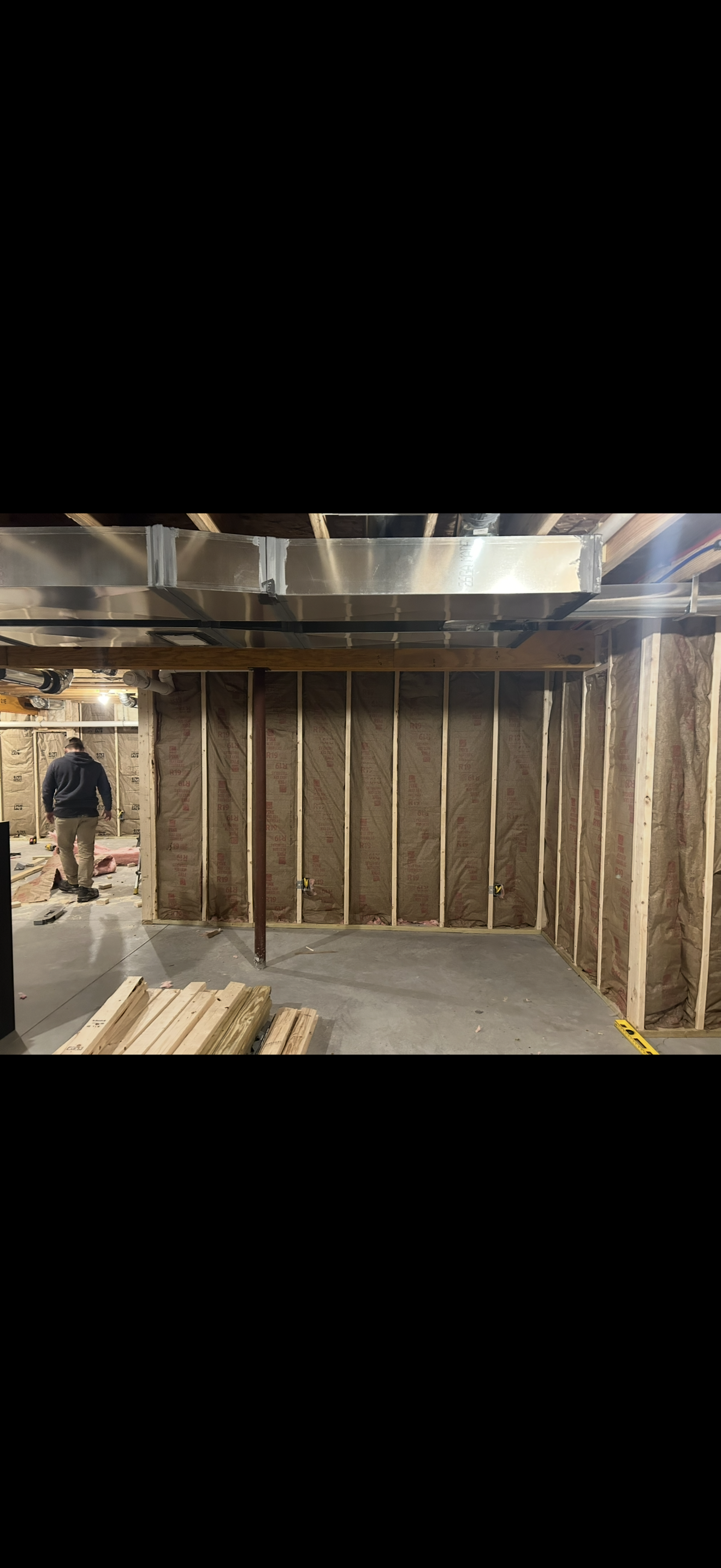 Interior of a room under construction with framing and insulation installed, a worker in the background, and construction materials on the floor.