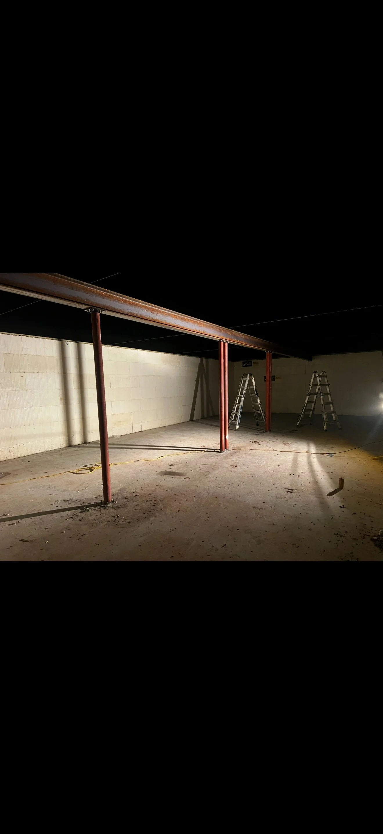 Interior of a building under construction with steel support beams, ladders, and construction equipment on a concrete floor.