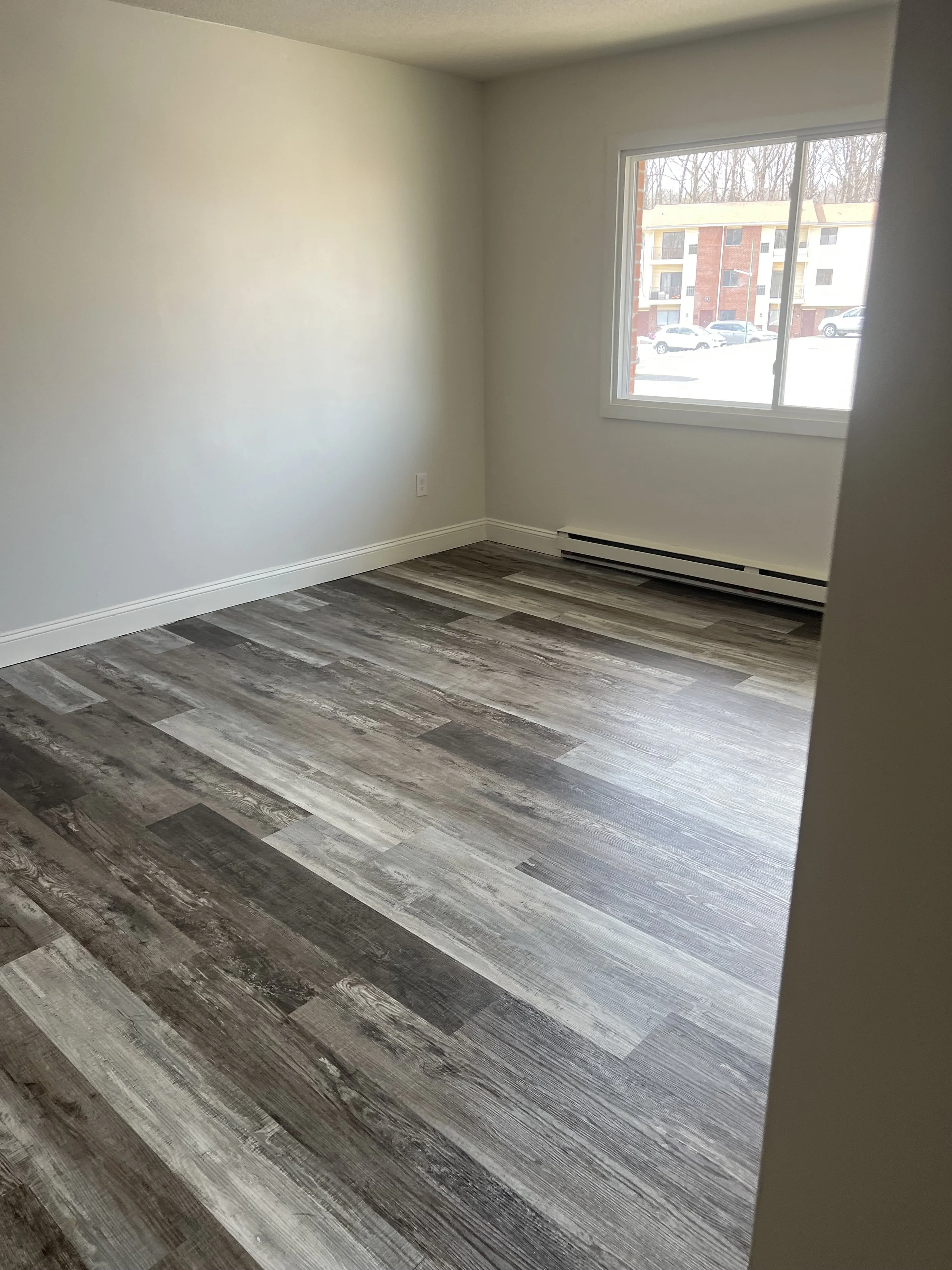 Empty room with light-colored walls, a large window showing an outside view of parked cars and apartment buildings, and wood-patterned flooring.