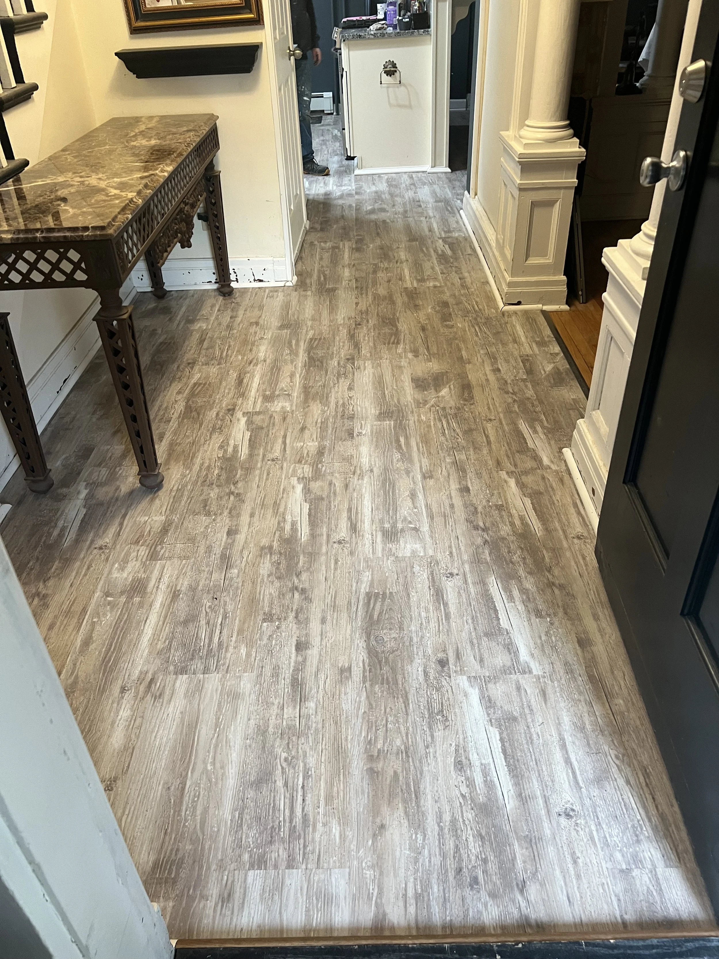View of a hallway with wood-look flooring, a table with a marble top on the left, and a black appliance door on the right. In the background, a person is partially visible near the kitchen doorway.