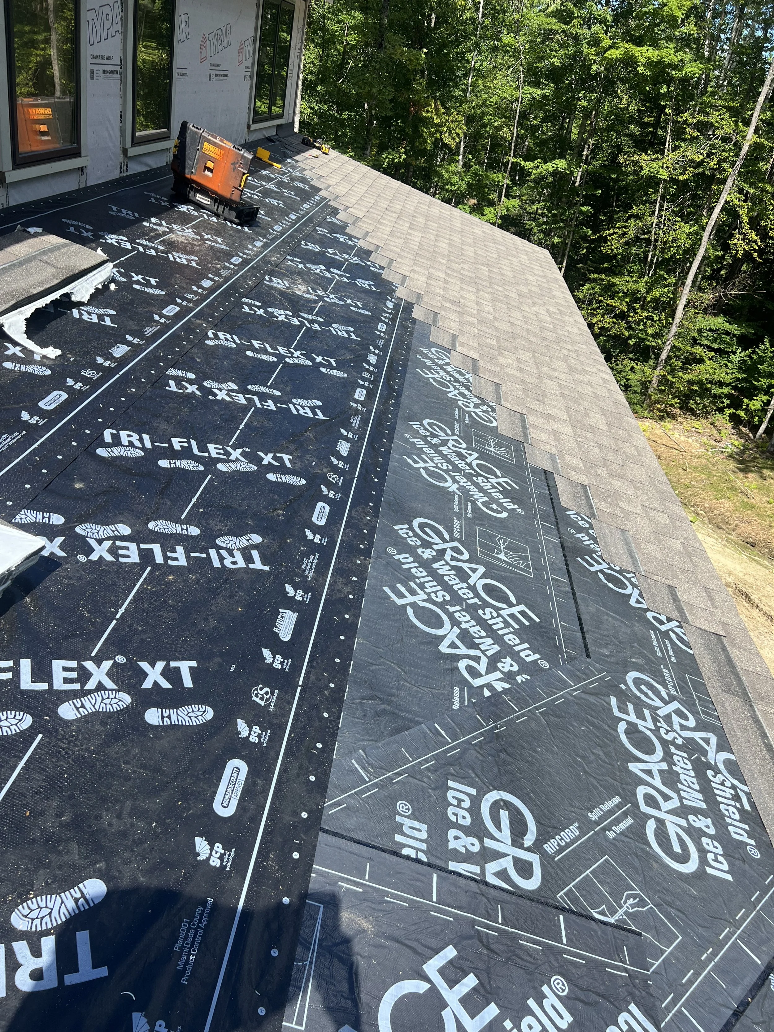 Roof under construction with black roofing underlayment labeled 'GRACE Ice & Water Shield' and new shingles partially installed, surrounded by trees.