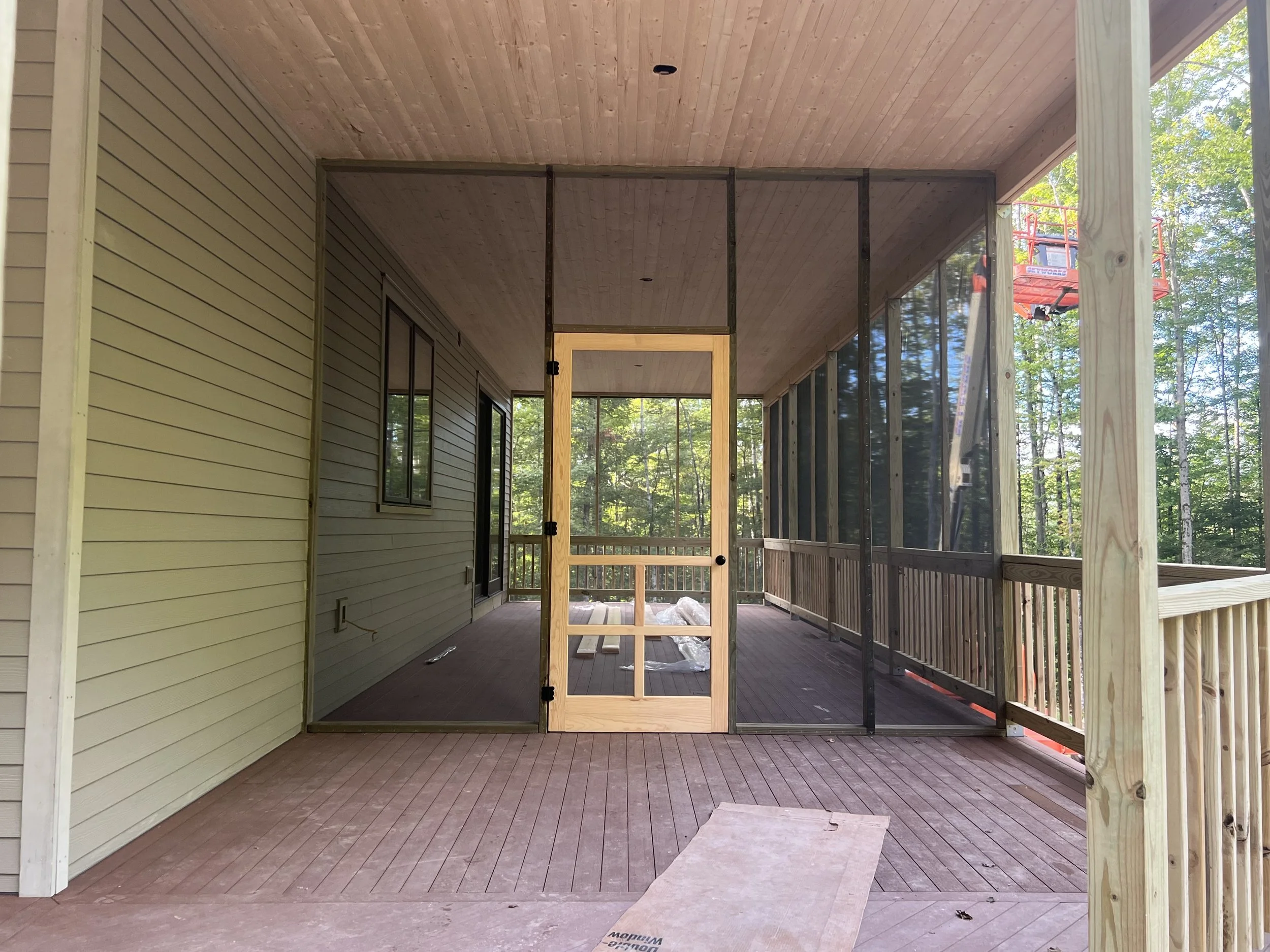 Under-construction screened porch or deck with partially finished framing, wood flooring, and adjacent house wall, surrounded by trees.