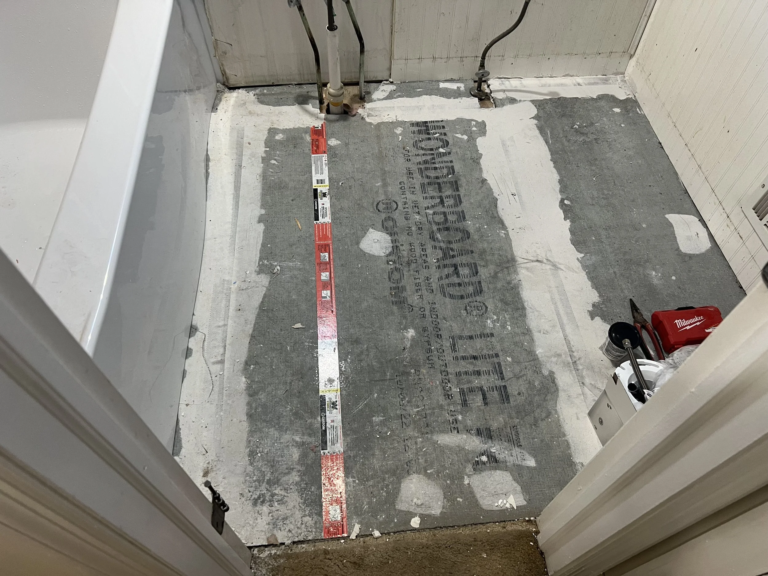Bathroom floor under renovation with exposed subflooring, a level placed on the floor, and tools including a container with a tool and a box with a power tool in the corner.