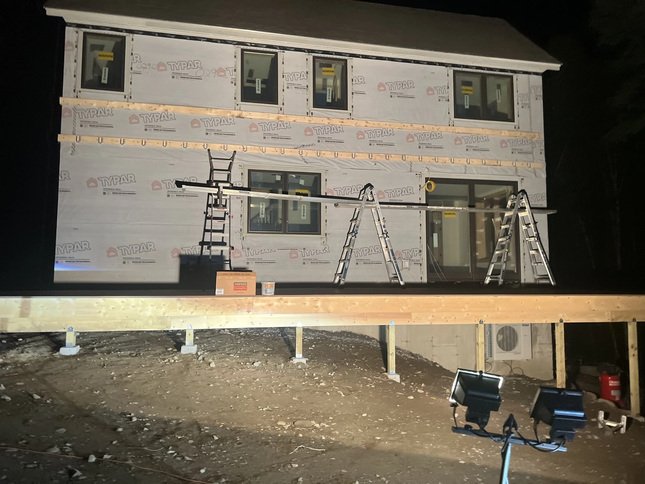 A residential house under construction at night, with completed upper and lower walls, multiple windows, ladders, construction tools, and bright floodlights illuminating the site.
