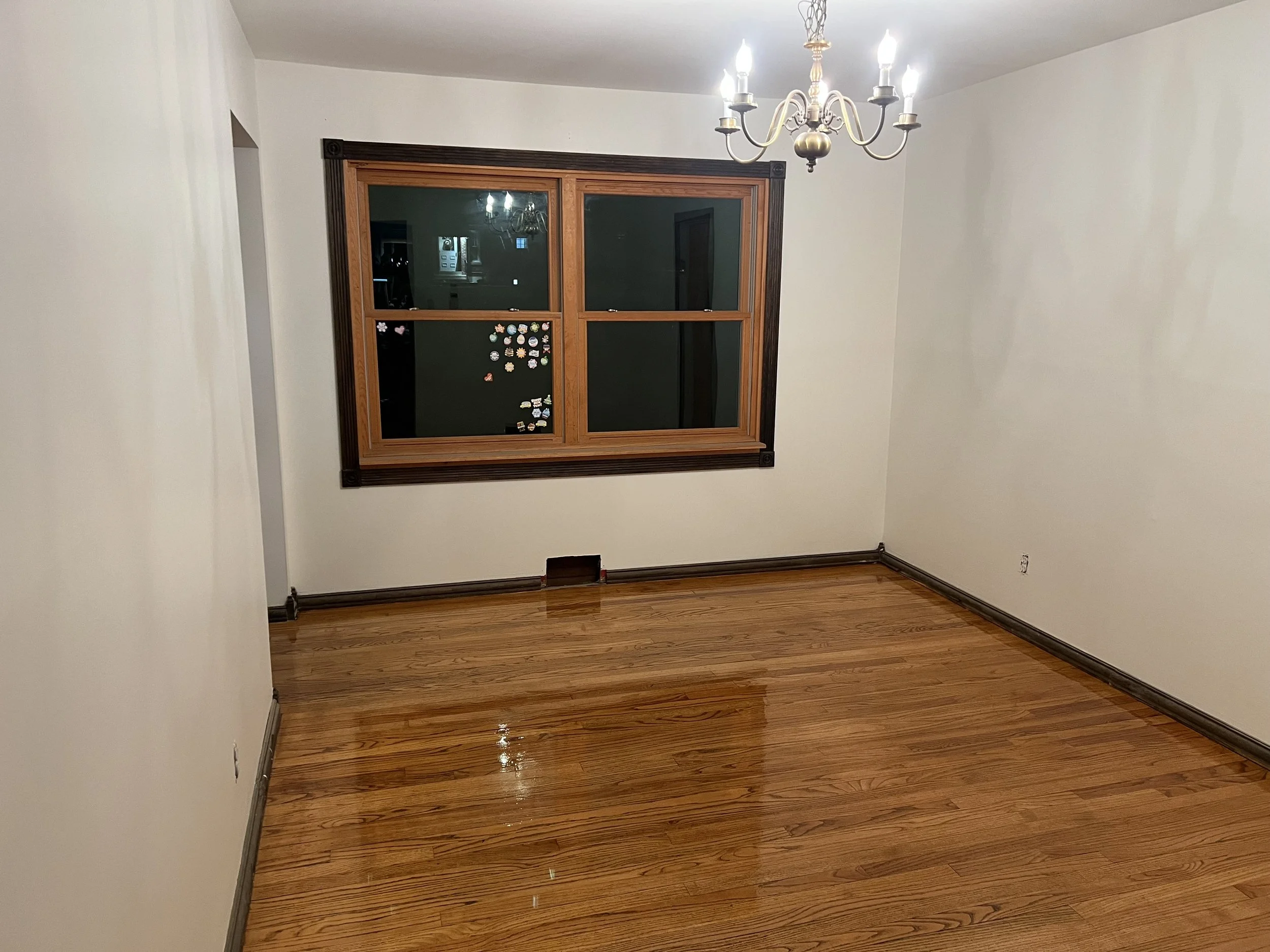 Empty room with hardwood floors, a window with a wooden frame, a white ceiling with an ornate chandelier, and white walls.