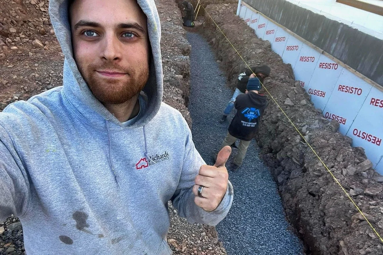 A man in a gray hoodie with a beard taking a selfie at a construction site, with two workers in the background working on a gravel pathway along a trench, surrounded by dirt and a wall with insulation panels marked 'RESISTO'.