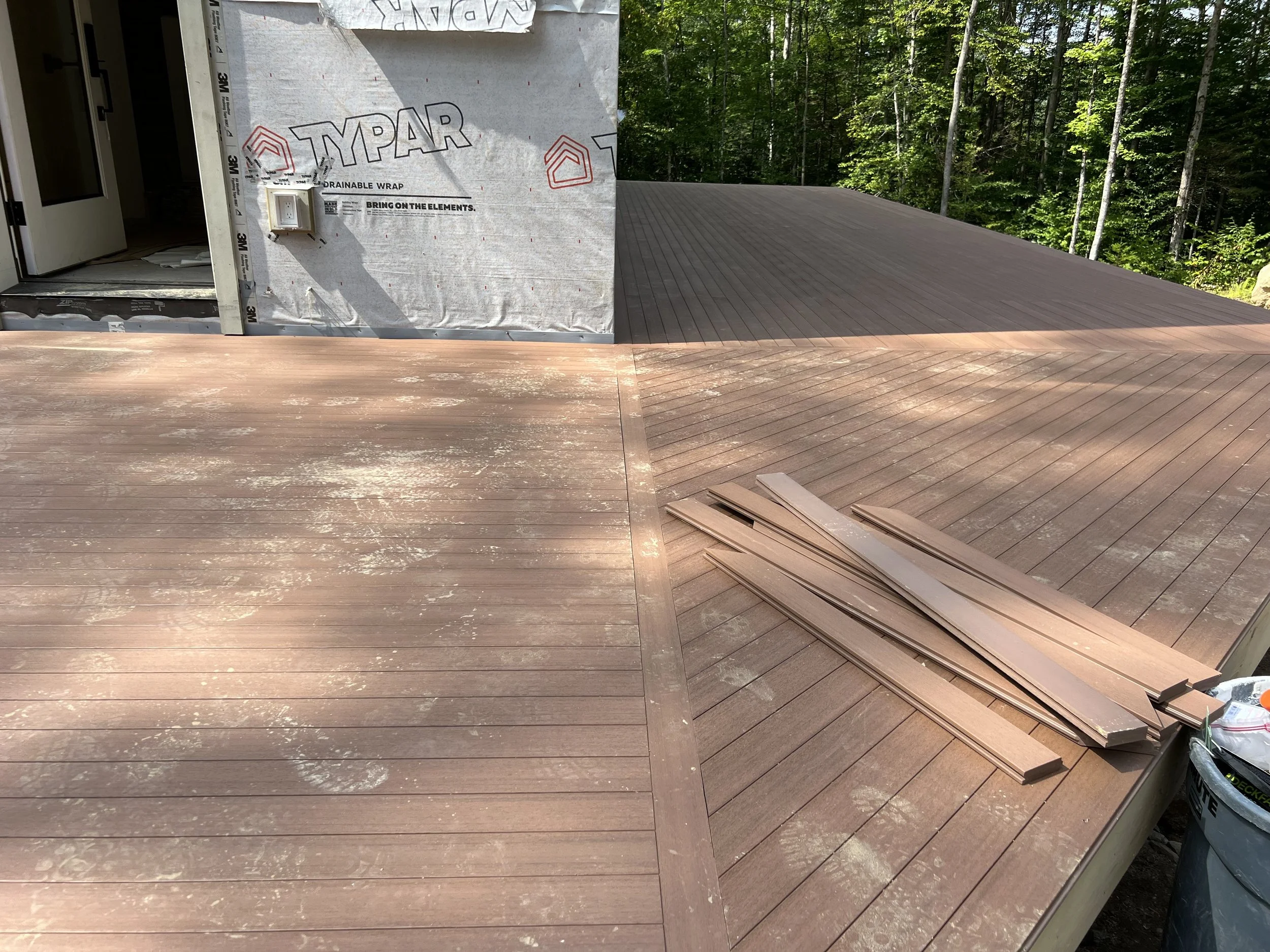 Newly installed wooden deck with some loose boards on the side, residential building under construction with Typar weather barrier and a forest in the background.