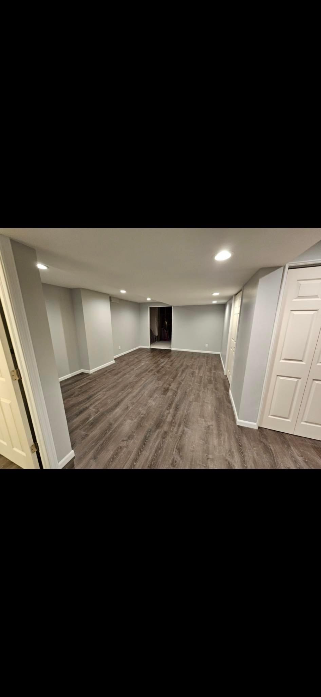 Empty room with light gray walls, dark wood flooring, and white trim, illuminated by recessed ceiling lights, with a closet door on the right.
