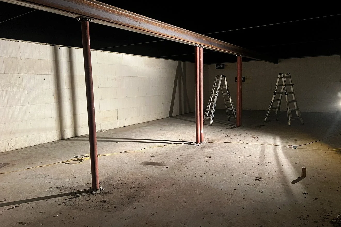 Empty underground parking garage with unfinished concrete floor, support beams, and two aluminum ladders leaning against the wall.