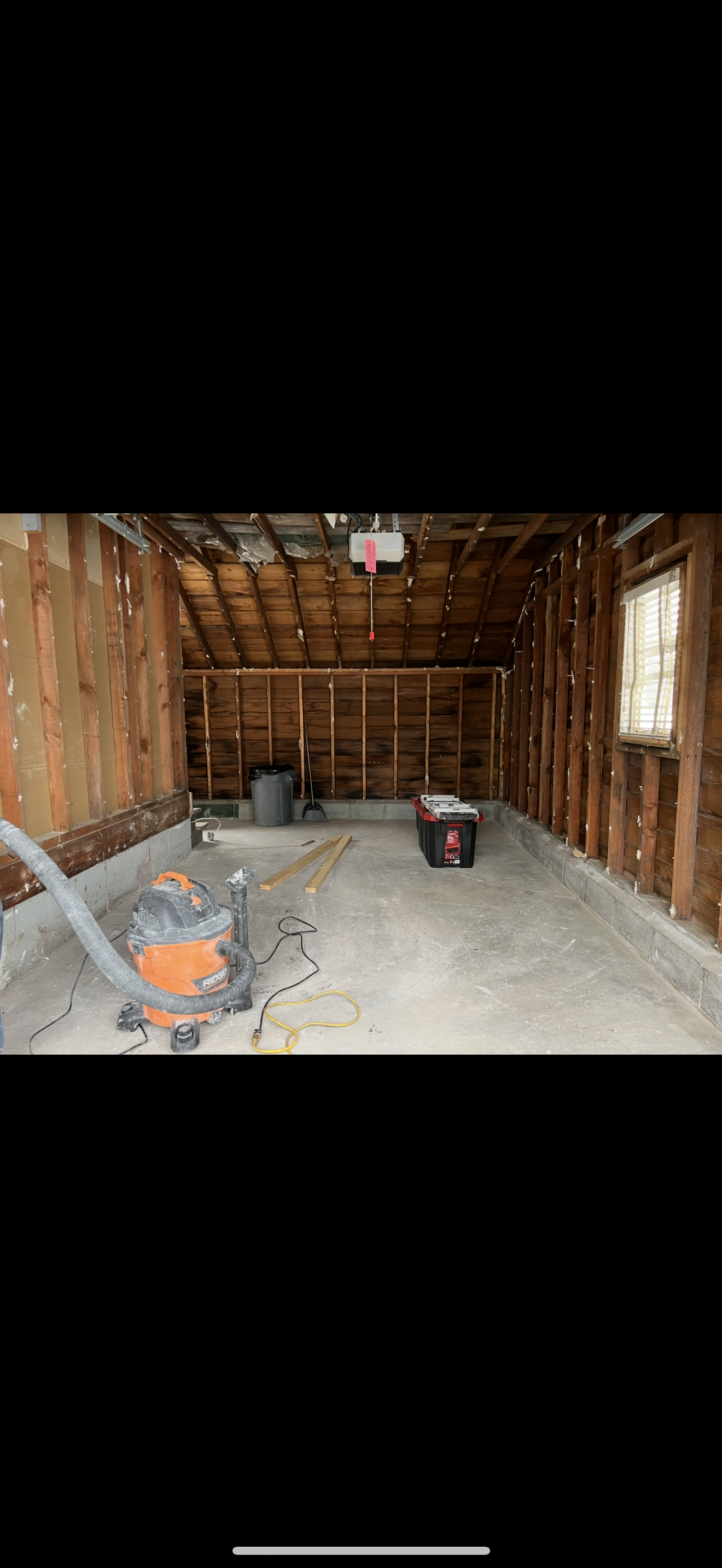 Interior of a garage under construction with exposed wooden framing, a concrete floor, a window, and construction tools including a wet/dry vacuum, a toolbox, and wooden planks.