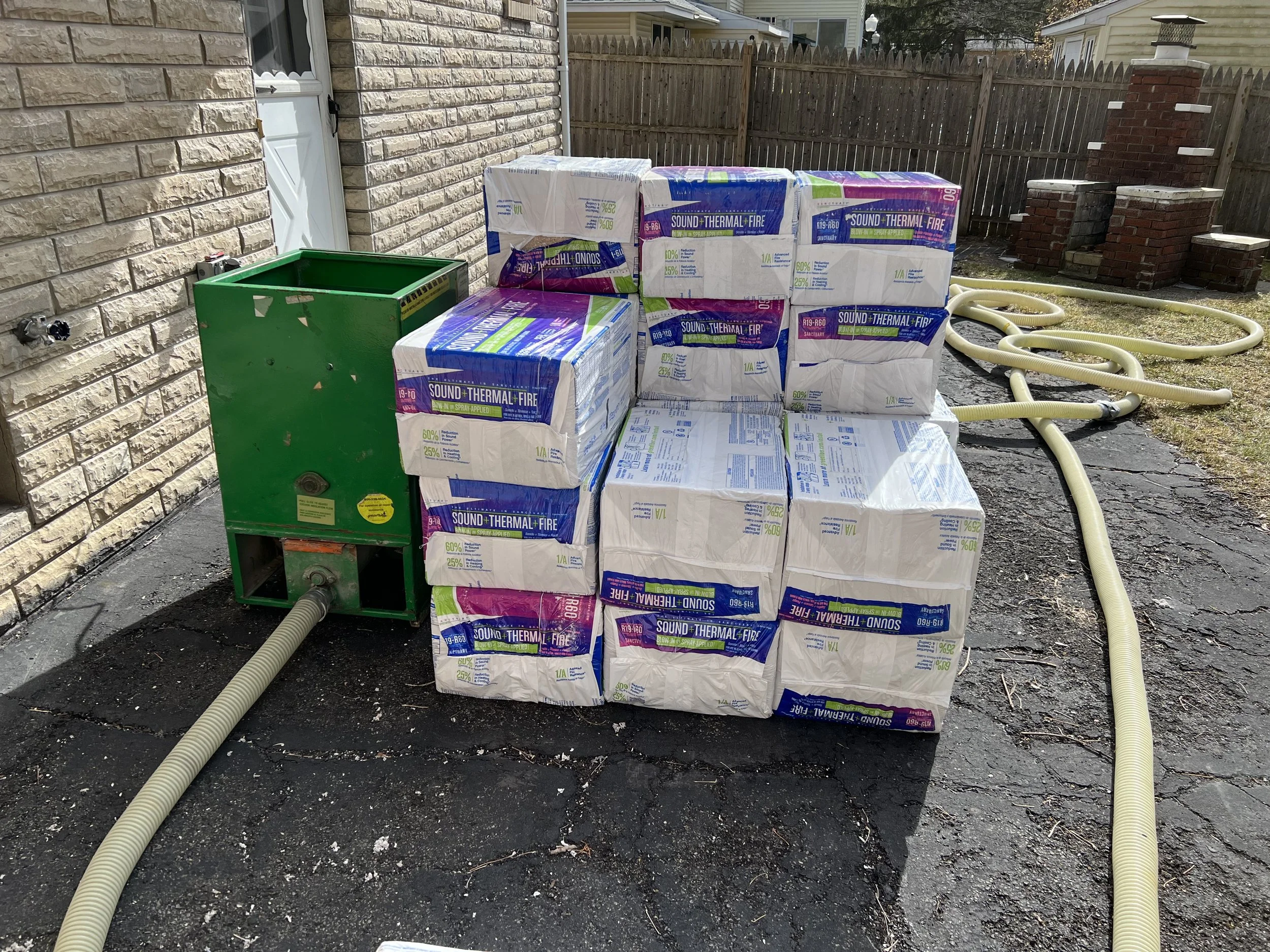 Piles of insulation material, labeled 'Sound Thermal Fire,' stacked outside on asphalt next to a brick house wall, with hoses and a green machine nearby.