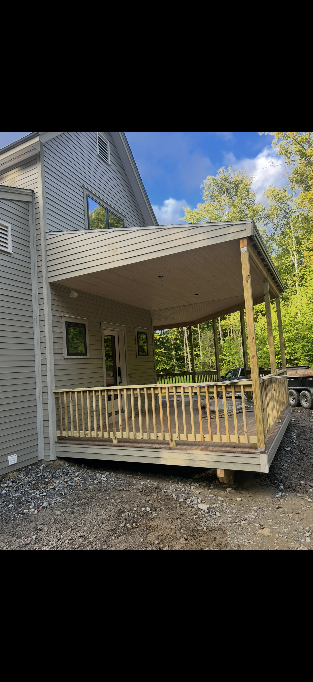 Newly constructed gray house with a covered wooden deck, surrounded by trees and a dirt ground.