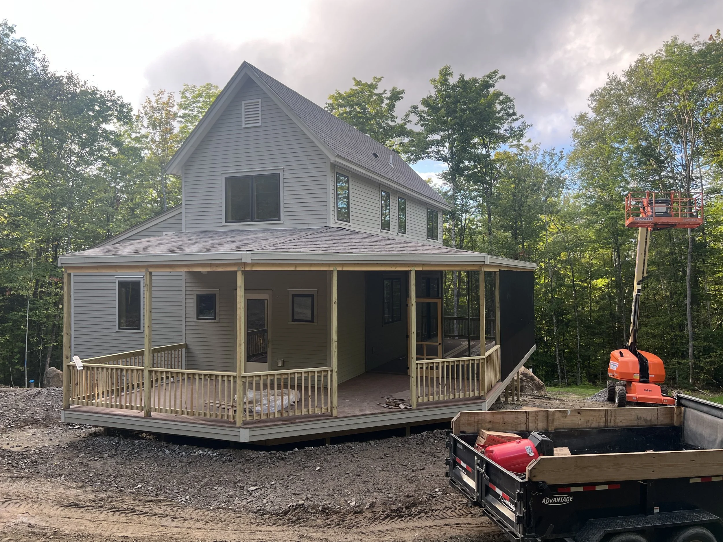 A house under construction with new wooden deck and railing, surrounded by trees, a cherry picker lift, and a trailer holding construction tools.