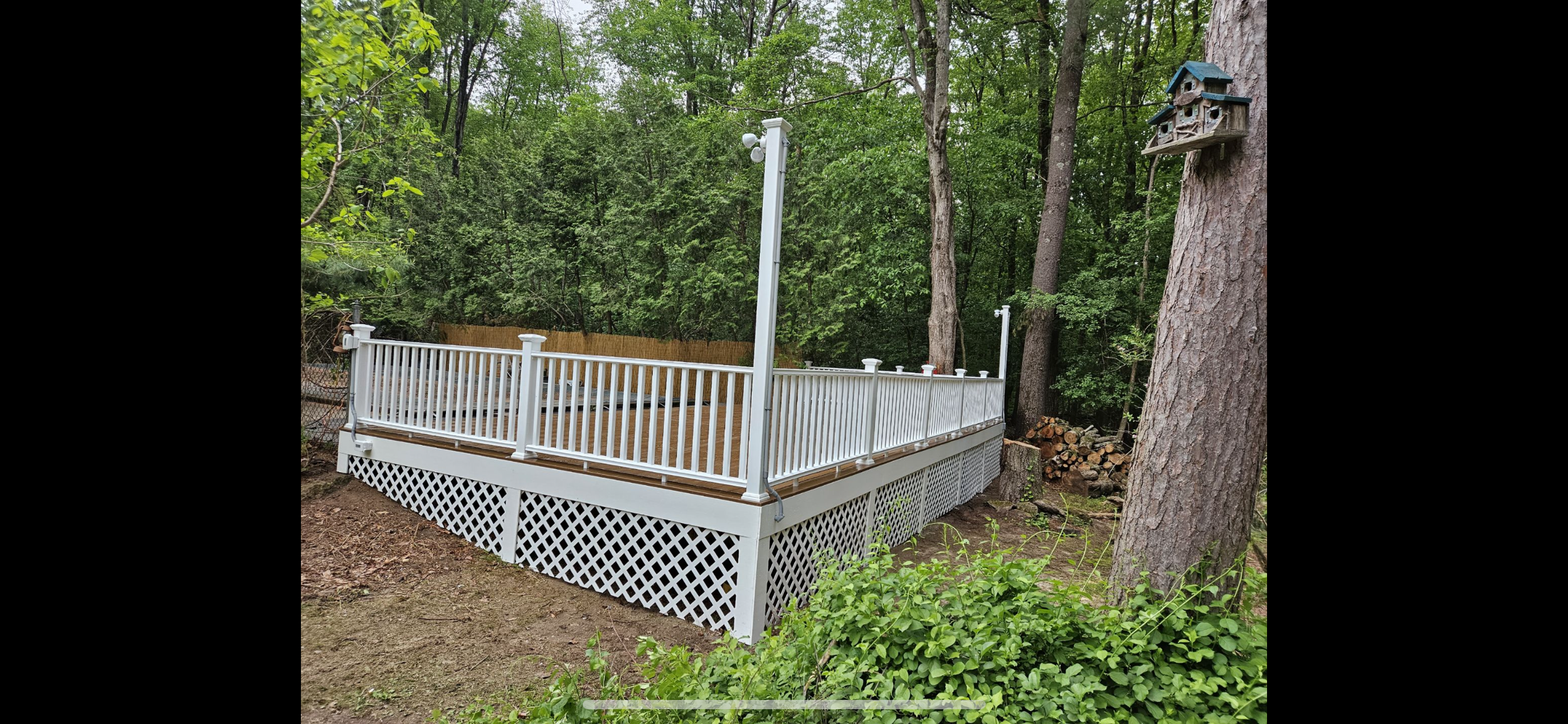 A new white wooden deck with a railing situated in a wooded backyard, with a wooden birdhouse mounted on a tree and firewood stacked nearby.
