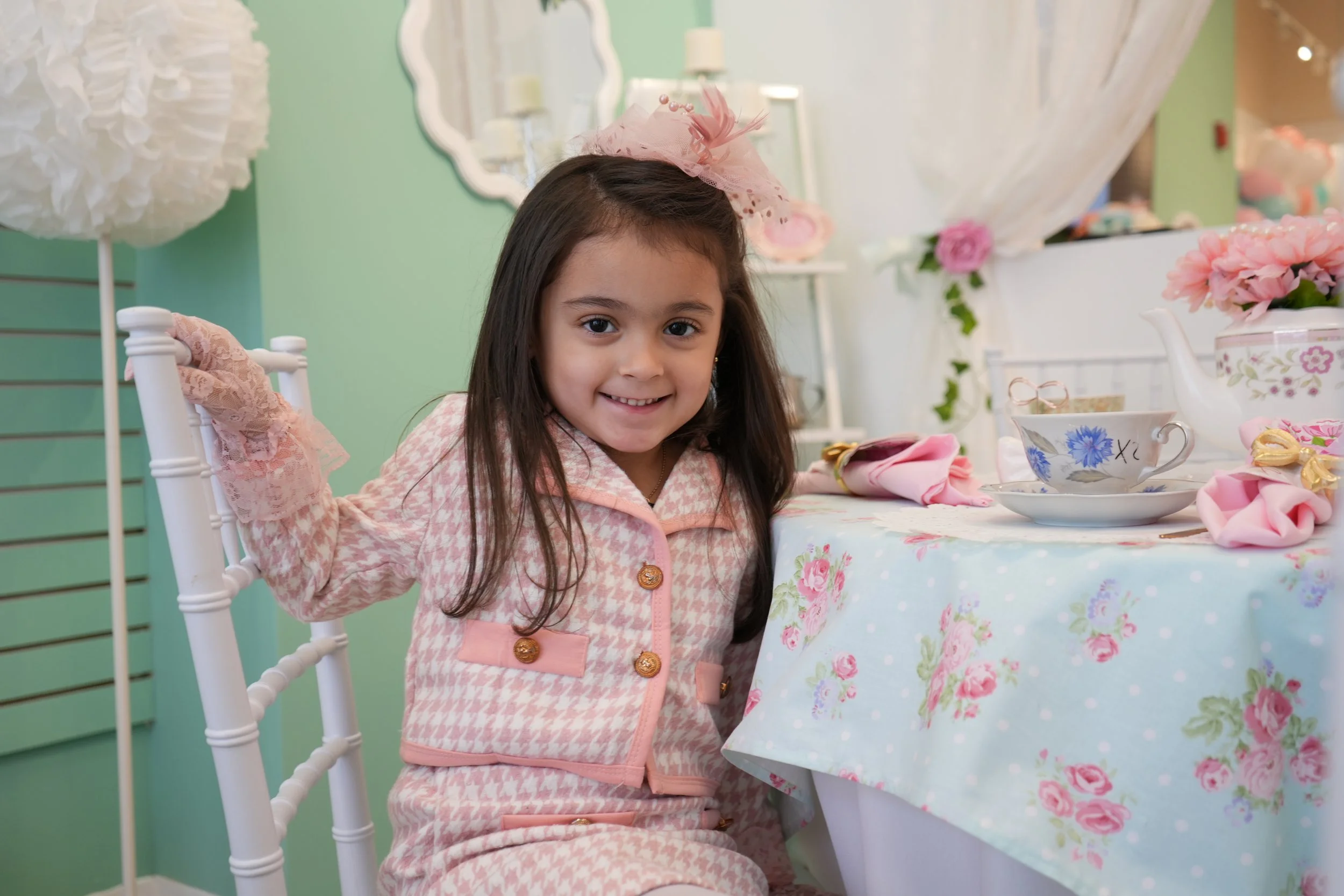 A table set for a celebration with a blue tablecloth, a white teacup with a gold trim on a matching saucer, a silver teapot, a spoon on a green napkin, and pearls, with a decorated background featuring balloons and streamers.