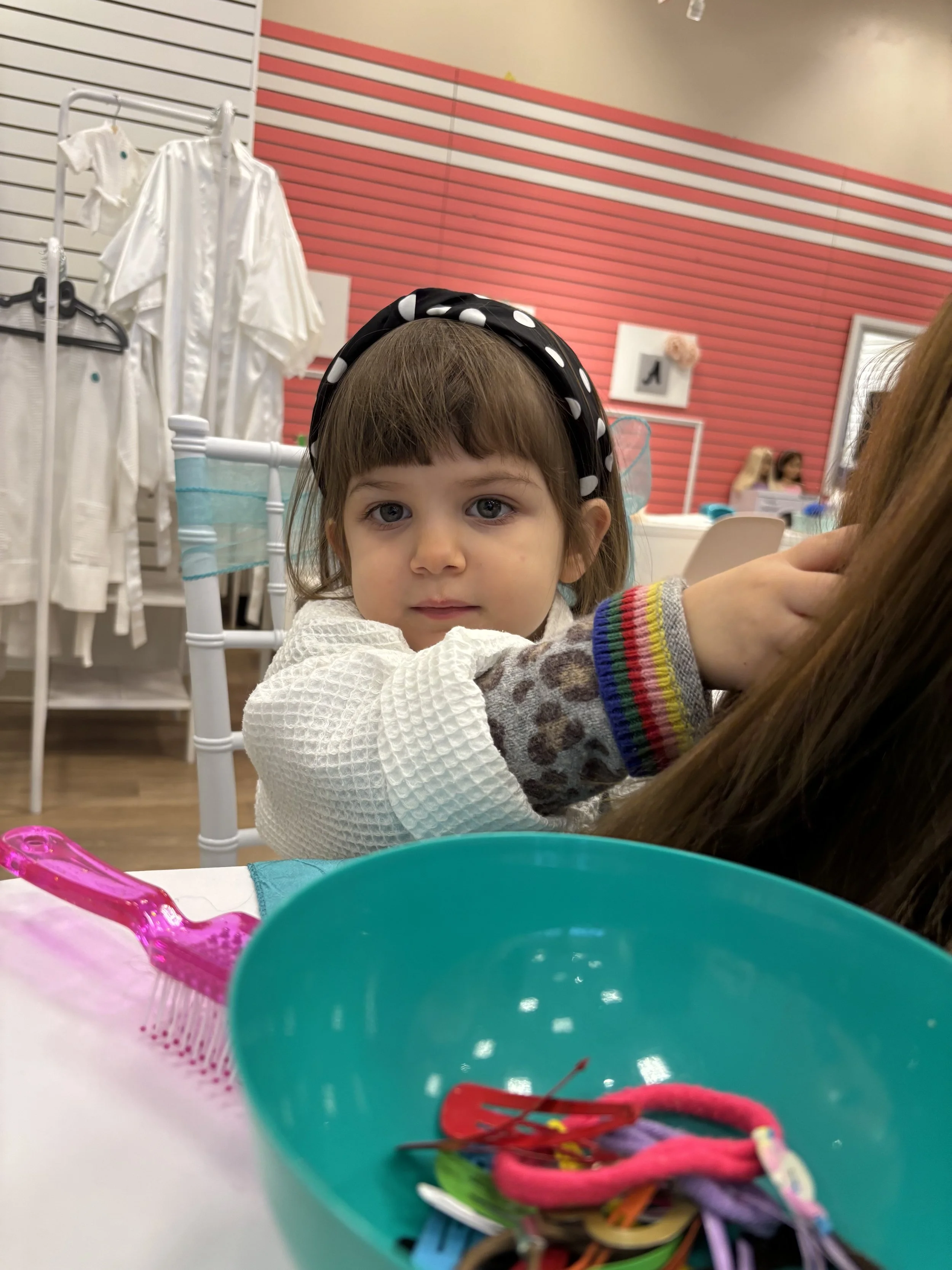 A young girl practicing hairstyling on a mannequin head at a craft table, wearing pink glasses, a wide-brimmed hat, and a pink vest, in a room decorated with balloon arrangements.