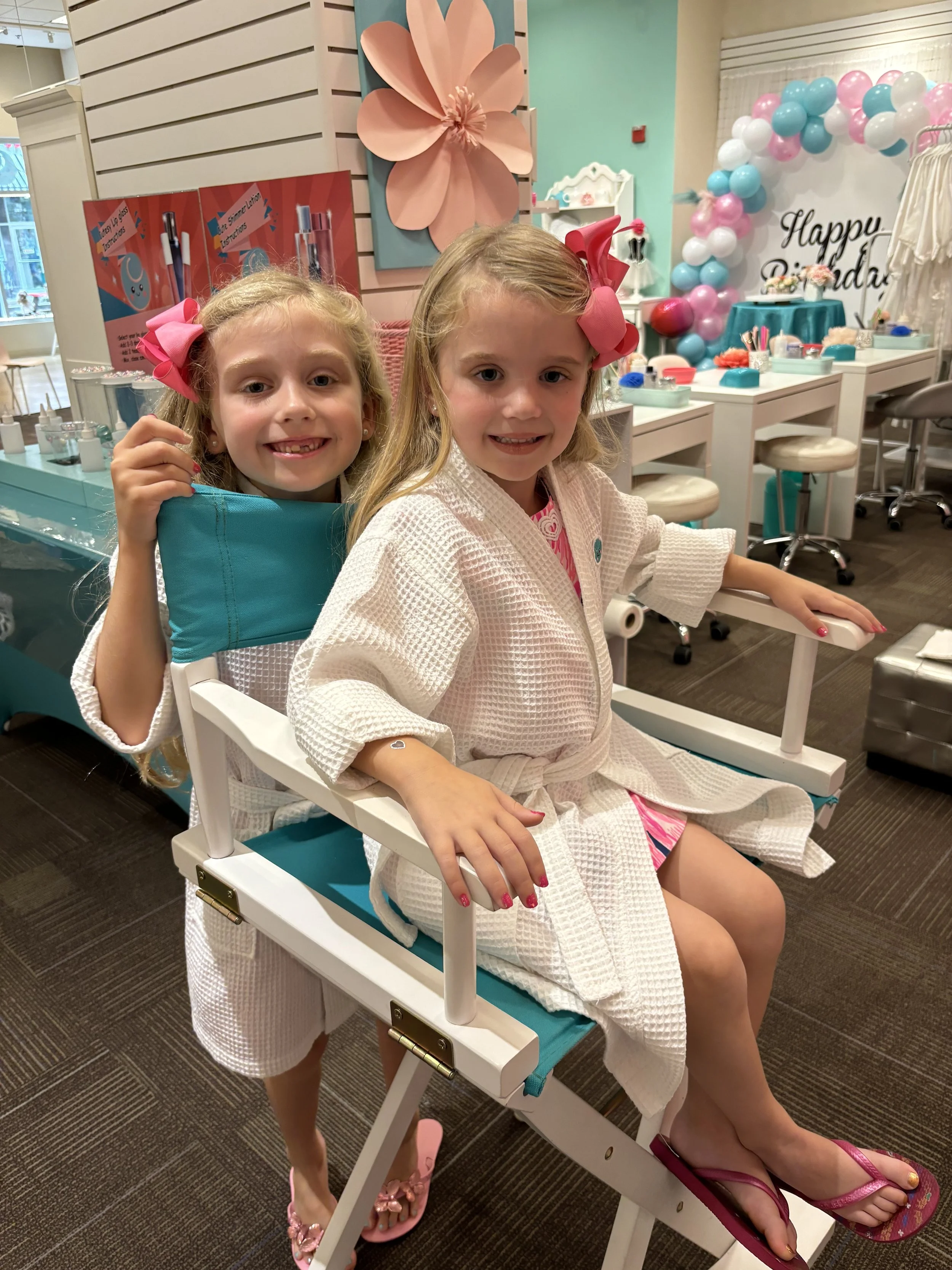 A young girl with a big blue bow in her hair sits at a nail salon table, getting her nails done by a person with bracelets. The table has nail polish bottles and tools, and there are other customers and mannequins with wigs in the background.