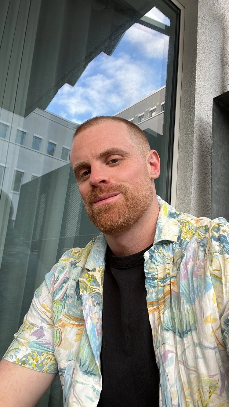 A young man with short hair and a red beard, wearing a colorful, subtle floral pattern shirt, sits in front of a large window through which you can see the blue sky with clouds.