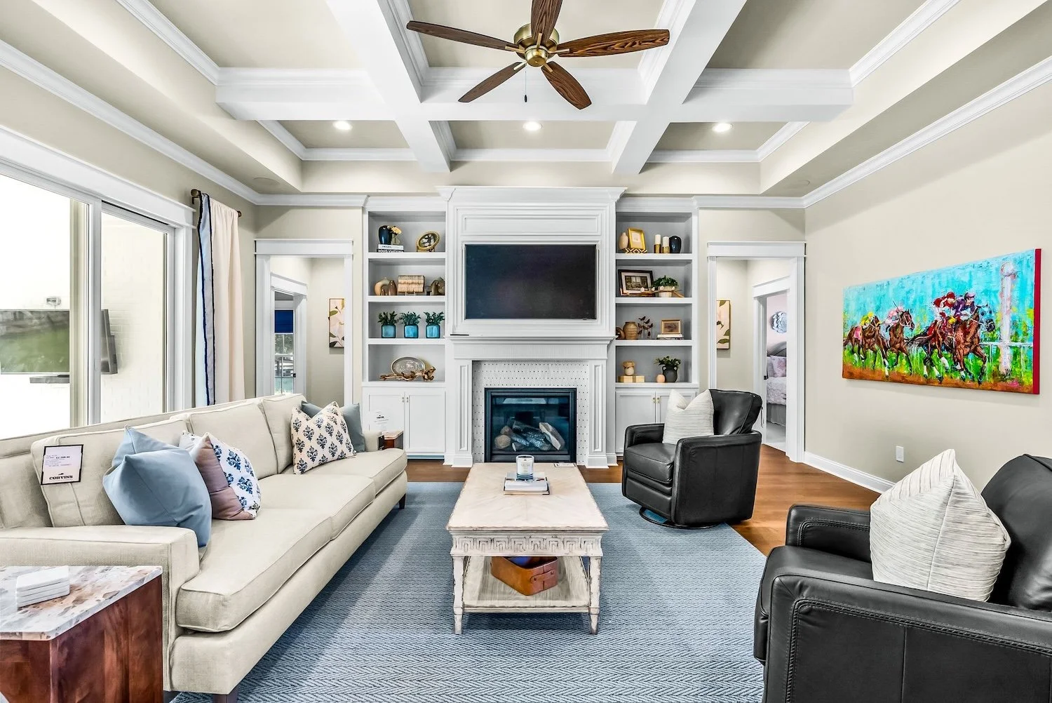 Living room with cream sofa, black armchair, white built-in shelves around a fireplace, a flat-screen TV above fireplace, colorful horse racing painting on wall, large windows with curtains, ceiling fan, hardwood floors, and decorative pillows.
