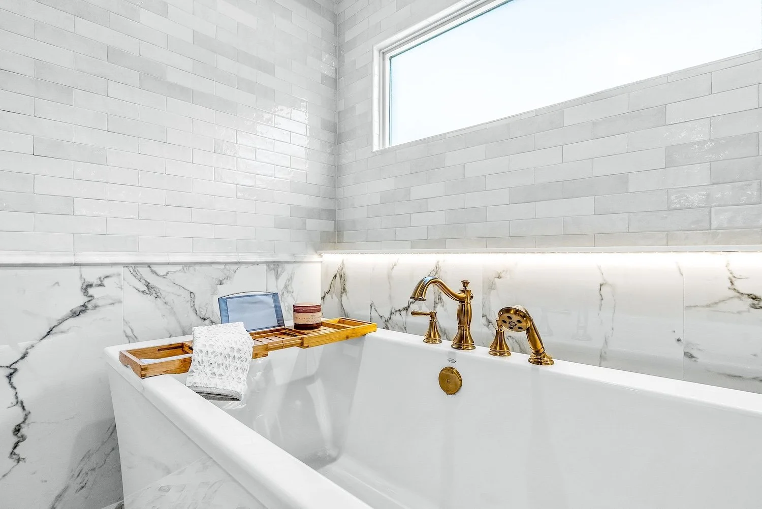 Modern bathroom with a white bathtub featuring gold fixtures, marble wall and a large window bringing in natural light.