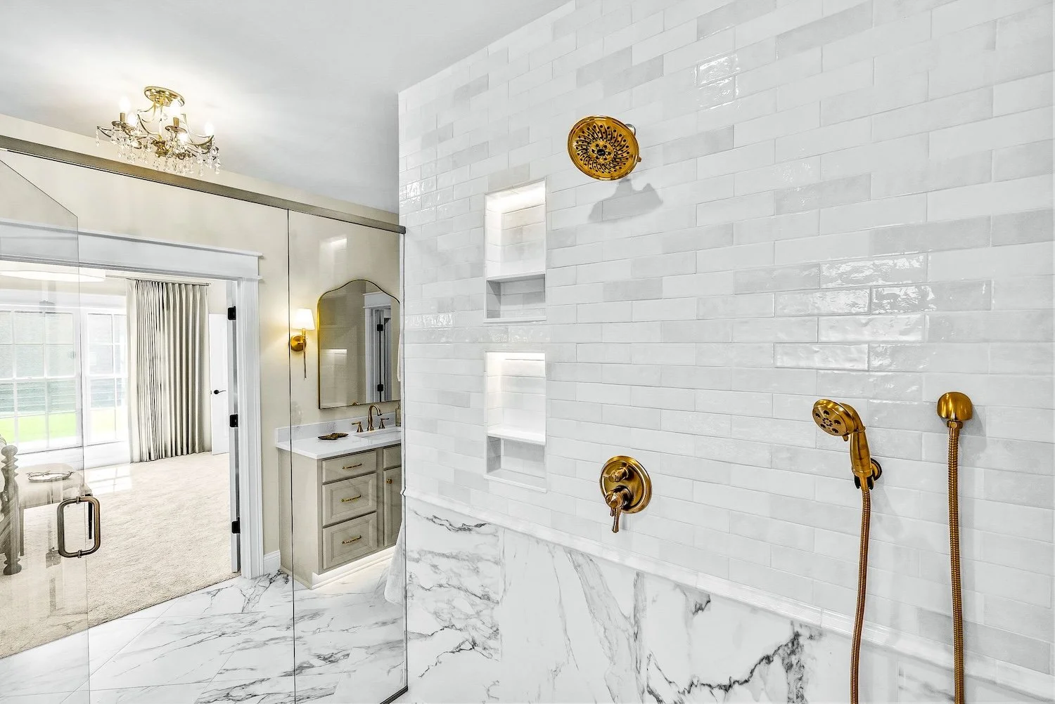 Bathroom walk-in shower with white subway tiles, gold fixtures, and marble flooring, with a view into a bedroom with a chandelier and sliding glass doors.