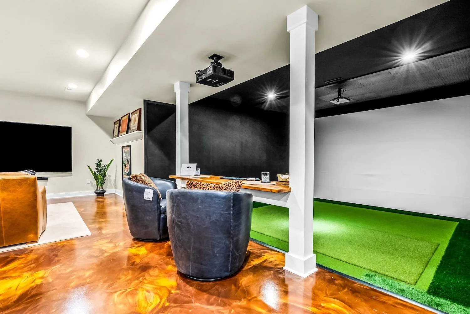A modern indoor lounge area with black leather chairs, a wooden table, a TV, and a green golf practice putting mat.