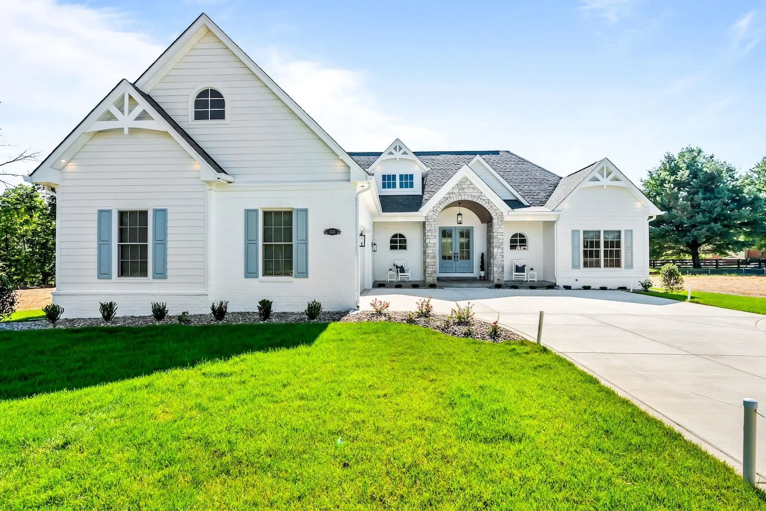 White modern house with gray shutters, stone archway, attached garage, green lawn, and a concrete driveway.