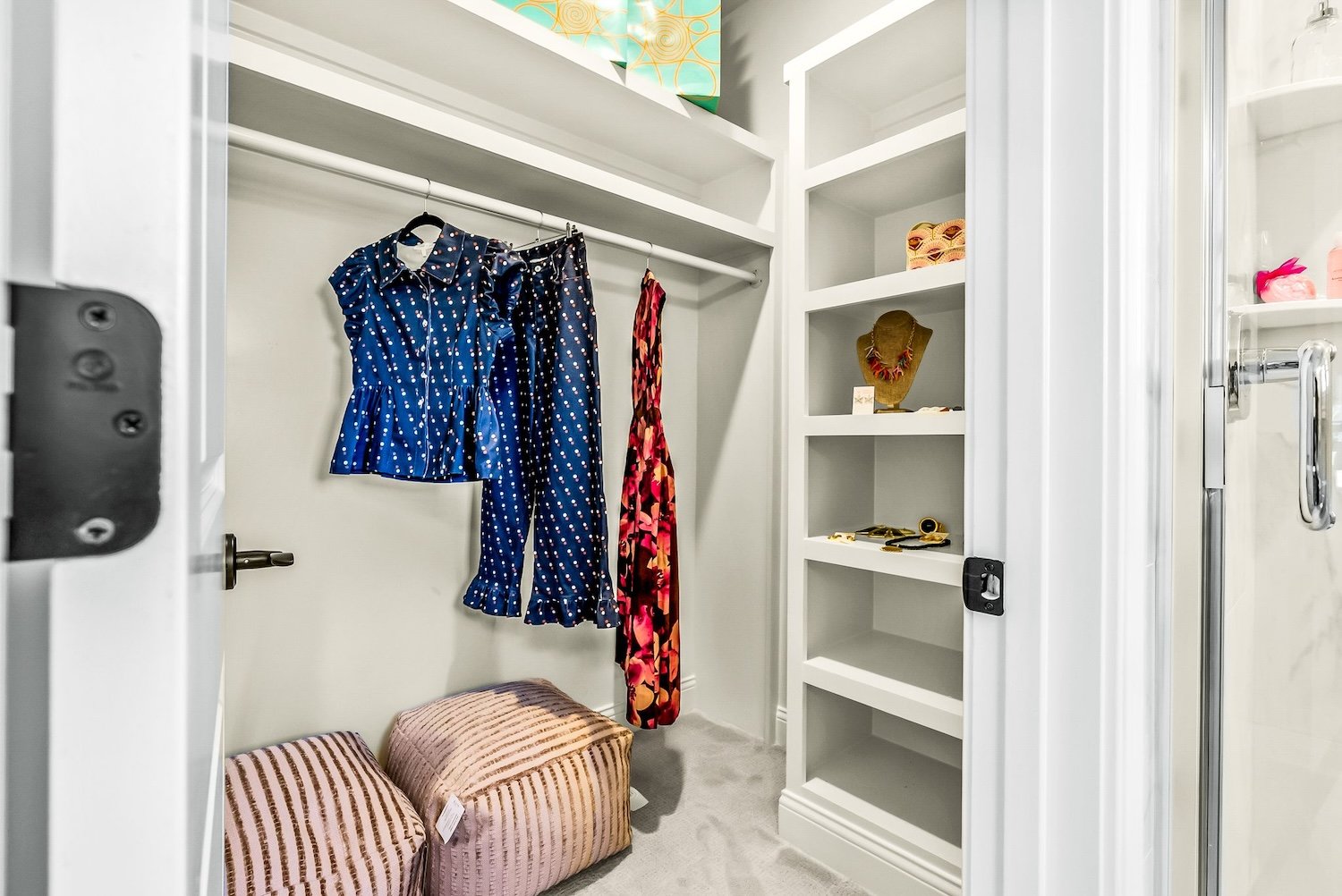 Inside a walk-in closet with hanging clothes, shelves with accessories, and two striped storage ottomans on a carpeted floor.