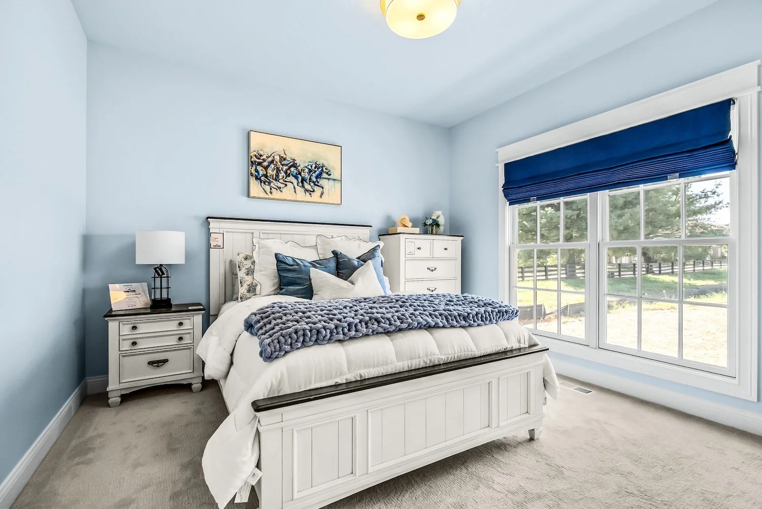 A bedroom with a white bed, blue pillows, gray knitted throw, nightstand with lamp, dresser with decorative items, large windows with blue blinds, and a painting of horses on the wall.