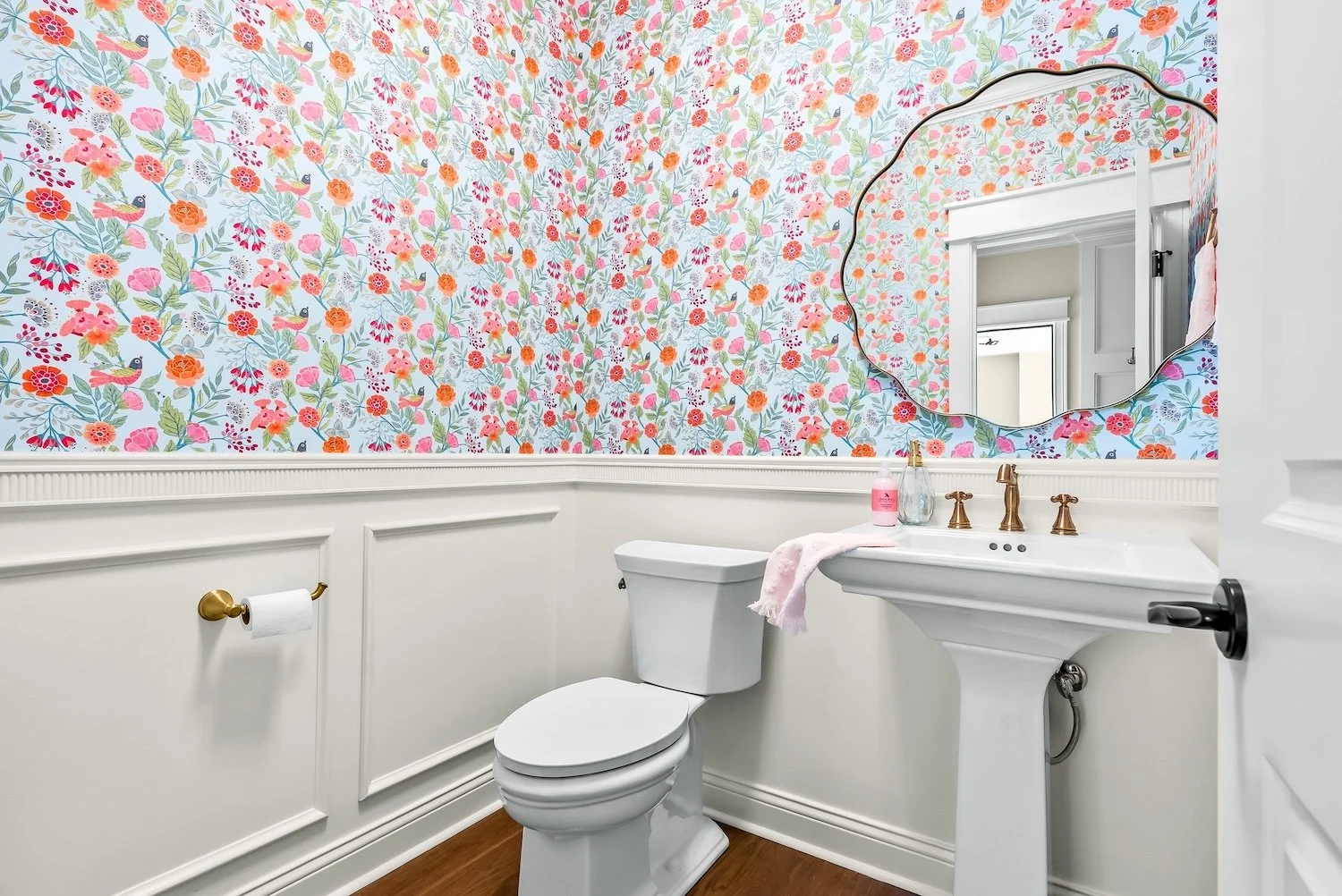 Bathroom with floral wallpaper, white toilet, white pedestal sink, oval mirror, brass fixtures, pink soap, pink towel, wood flooring, wall molding, and window reflected in the mirror.