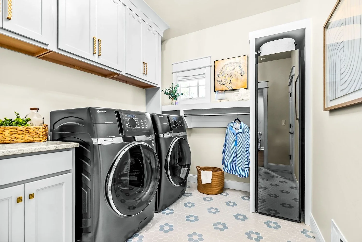 A laundry room with white cabinets, a marble countertop, two black front-loading washing machines, a basket and a laundry basket, a hanging blue and white striped shirt, a small window, a mirror, and wall art.