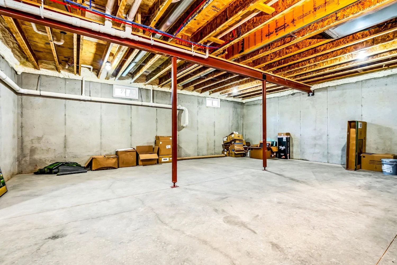Empty basement with concrete walls and floor, exposed ceiling with wooden joists and pipes, and some boxes and items stacked along the walls.