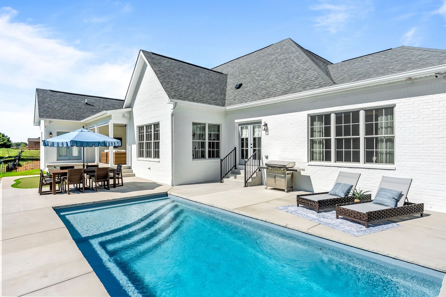 Backyard with a swimming pool, outdoor furniture including a dining table with an umbrella, two lounge chairs with pillows, a grill, and a white house with large windows and a door with stairs.