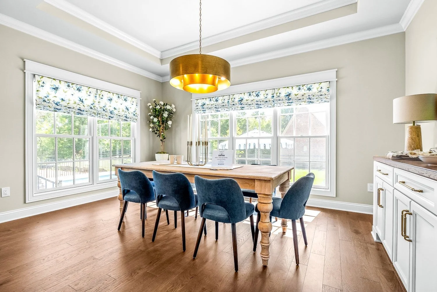 Bright dining room with large windows, a wooden table, six blue upholstered chairs, a white sideboard, and a modern gold pendant light fixture.