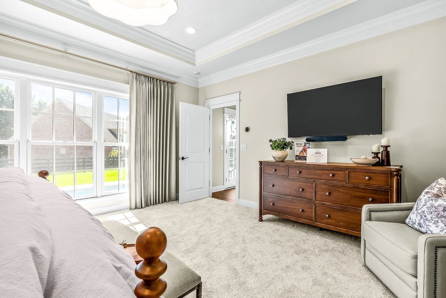 A cozy bedroom with a large window, cream-colored walls, beige carpet, a wooden dresser, a wall-mounted TV, a white armchair with a decorative pillow, and a door leading to a balcony with a view of neighboring houses.