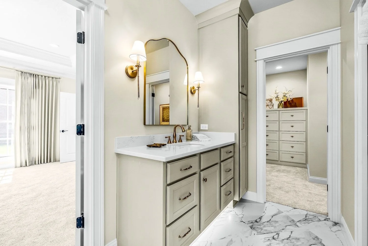 Bathroom with beige walls, a mirror, two wall sconces, a beige vanity with brass handles, a white marble countertop, a gold faucet, and a small decorative tray. Adjacent to the bathroom is a walk-in closet with built-in shelving and drawers, carpeted