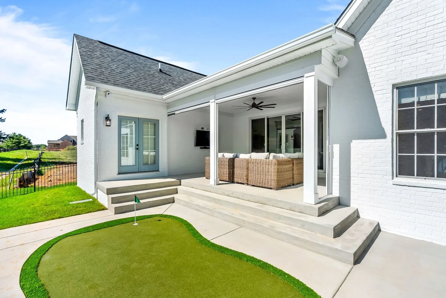 Backyard patio of a white house with a screened-in porch, outdoor seating, a ceiling fan, and a small putting green with a flag, green grass, and steps leading down to a concrete walkway under a blue sky.