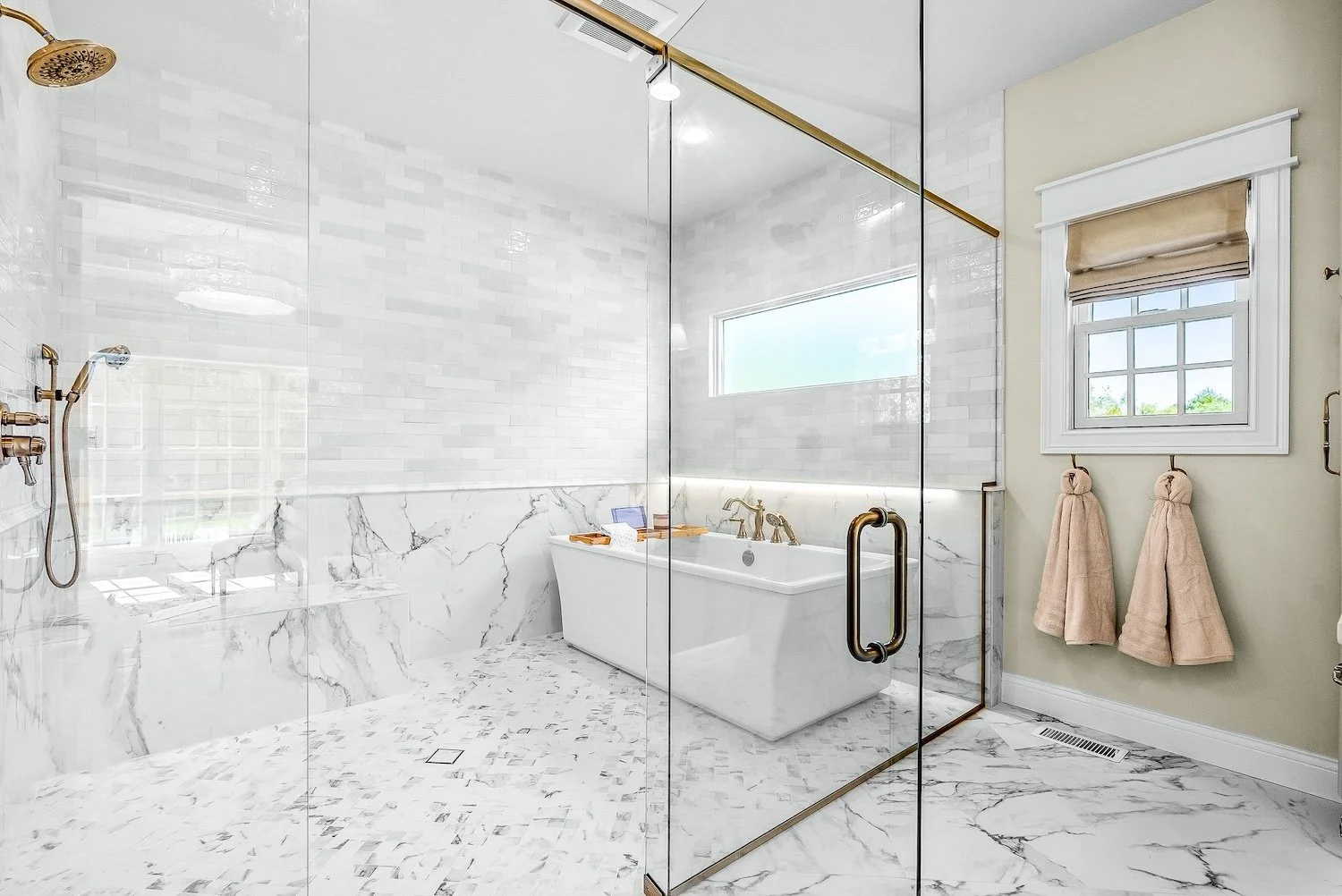 Modern bathroom with a glass shower enclosure, a white bathtub with gold fixtures, a window, and beige towels hanging on a wall.
